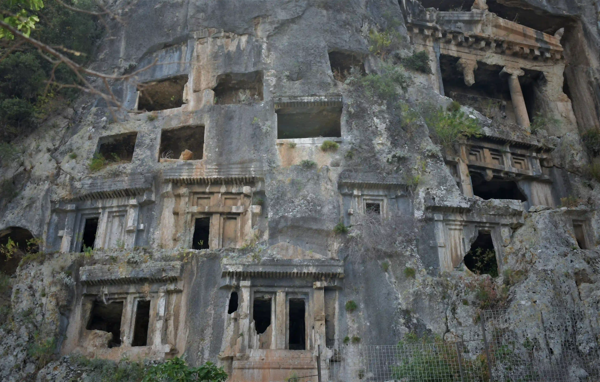 Ancient rock-cut tombs with temple-like facades carved directly into a steep, rugged stone cliff.