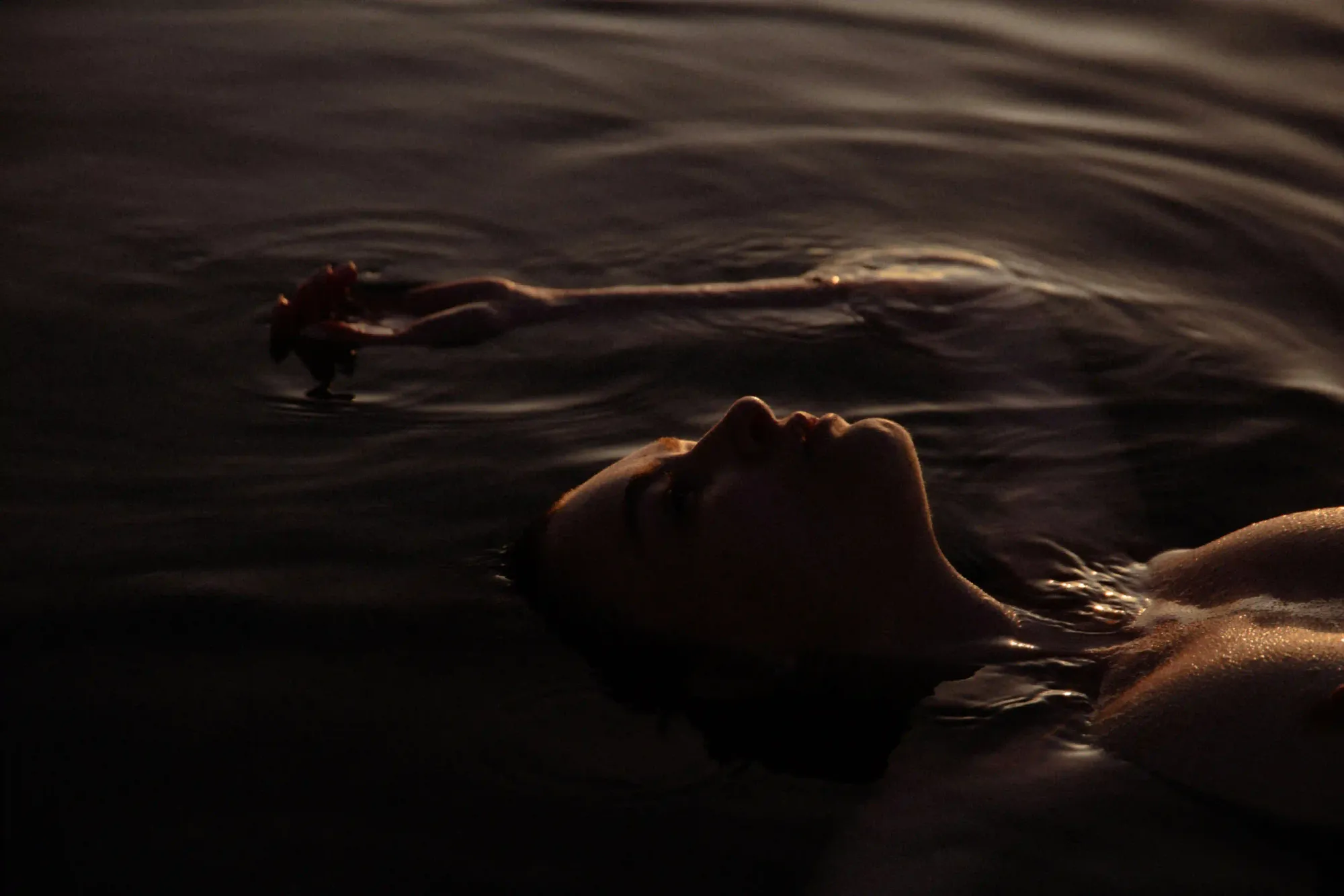A serene close-up of a person floating peacefully on their back in dark, calm water at dusk, representing the deeply restorative state of the blue mind.