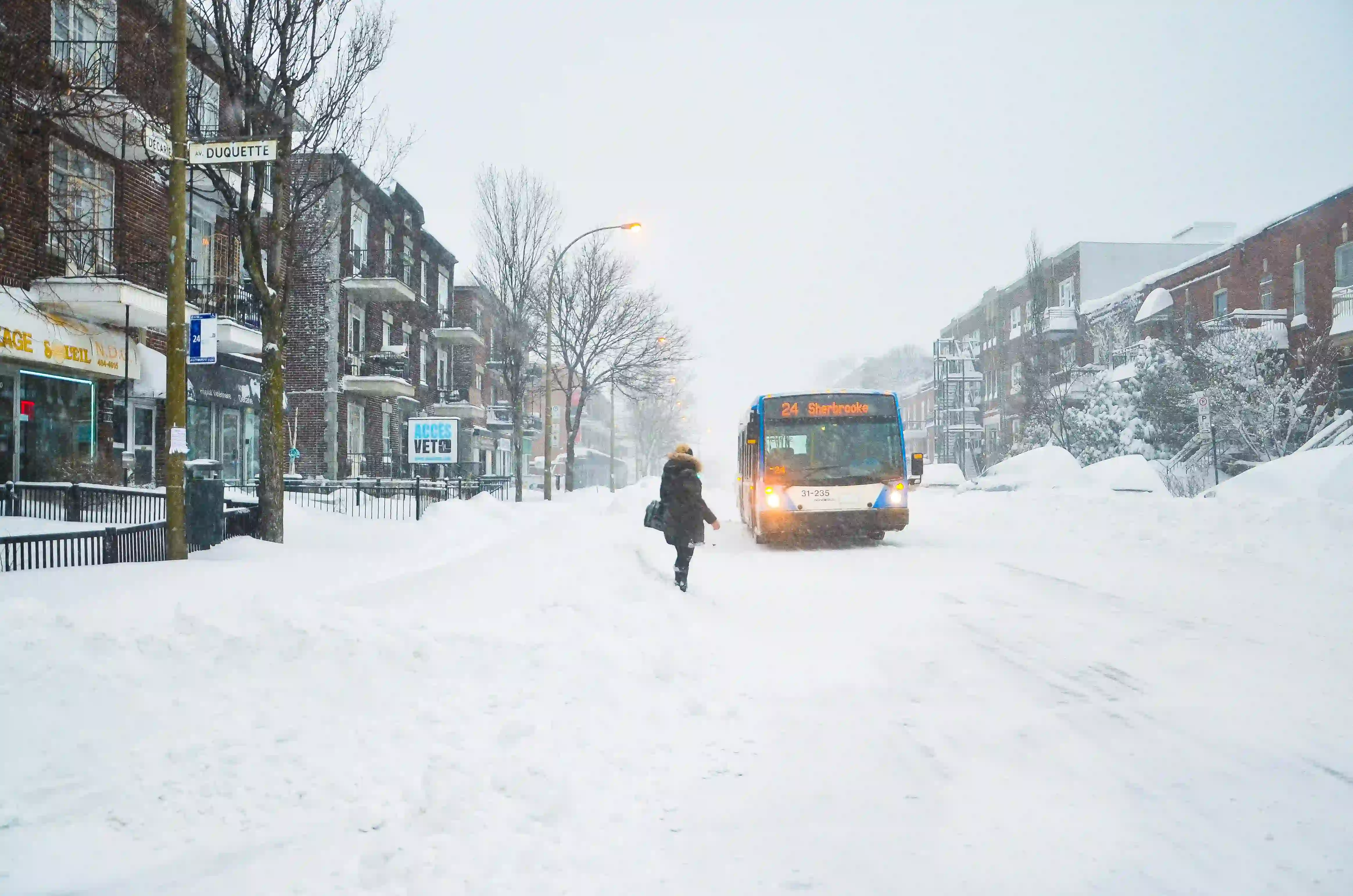 A lone pedestrian walking towards a city bus in the middle of a heavily snow-covered street during a whiteout winter storm in Montreal.