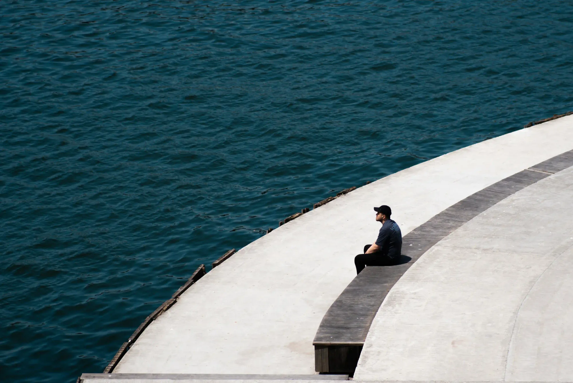 A lone man wearing a black cap sitting on a curved concrete and wooden promenade, looking out at the deep blue water.