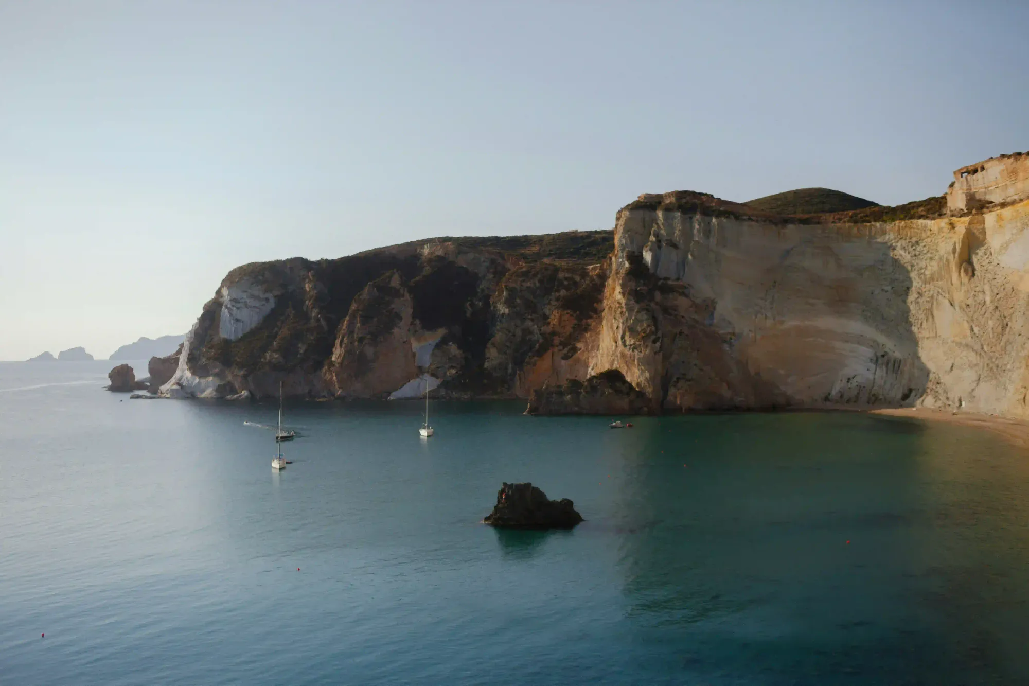 A wide, calm Mediterranean bay surrounded by steep, rugged cliffs under a slightly hazy, faded sky, with a few sailboats anchored in the water.
