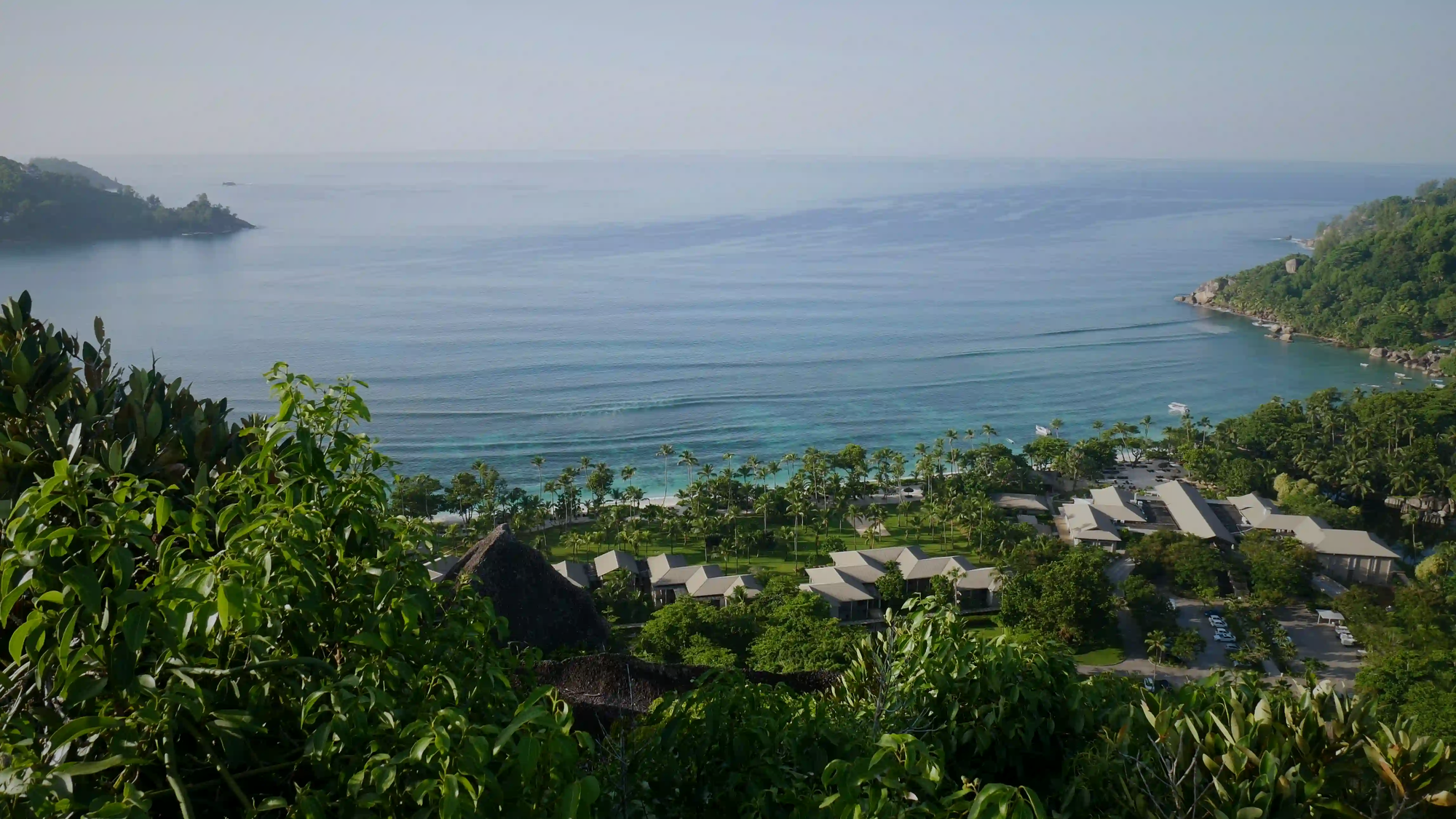 A secluded coastal retreat nestled within lush green foliage, overlooking a tranquil blue bay. The architecture blends seamlessly into the natural landscape, offering a silent, neuro-restorative escape from the modern world.