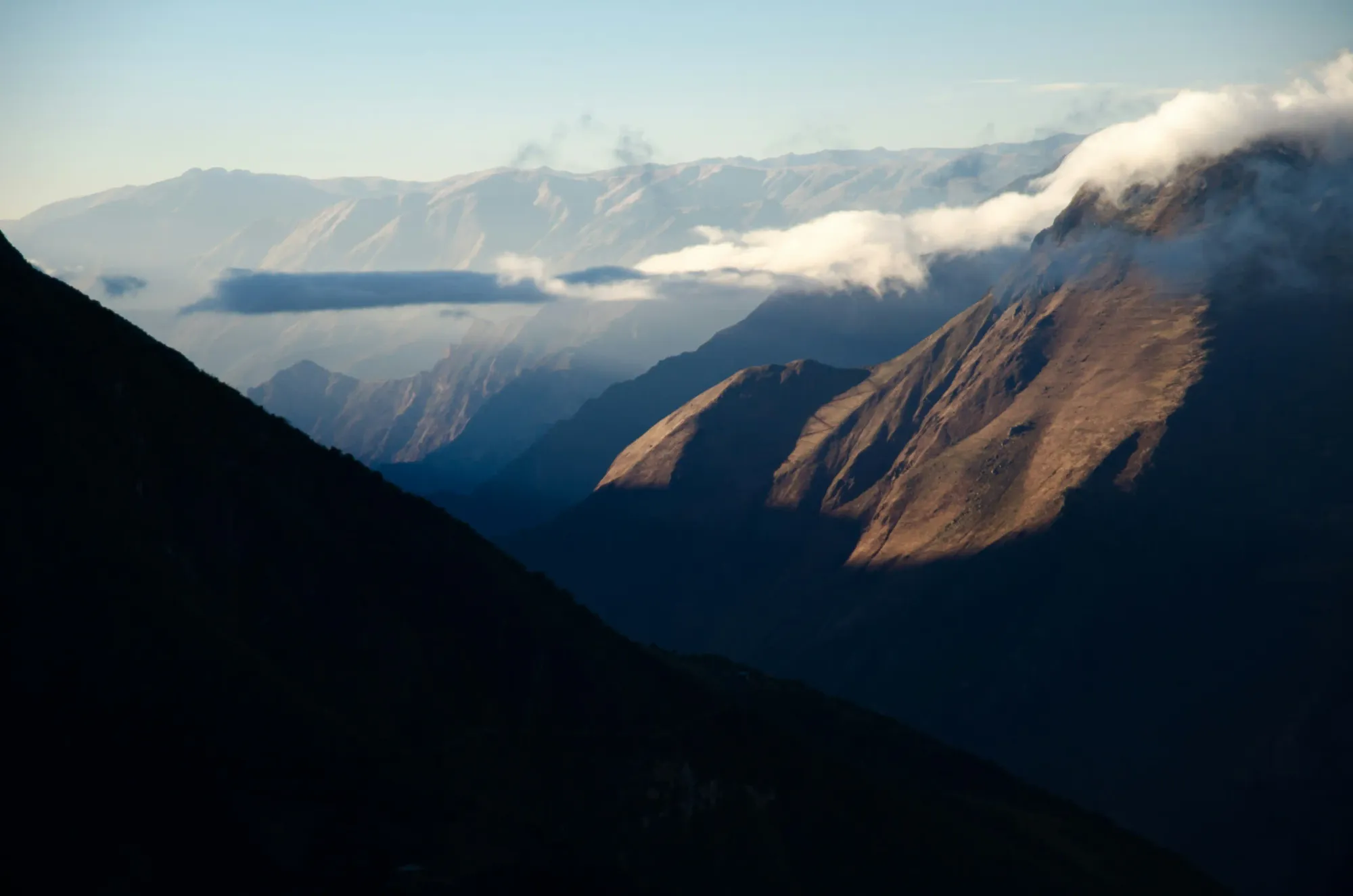 Layered mountain peaks of the Peruvian Andes bathed in soft golden sunlight, with deep shadows in the foreground and thin clouds passing through the vast valley.