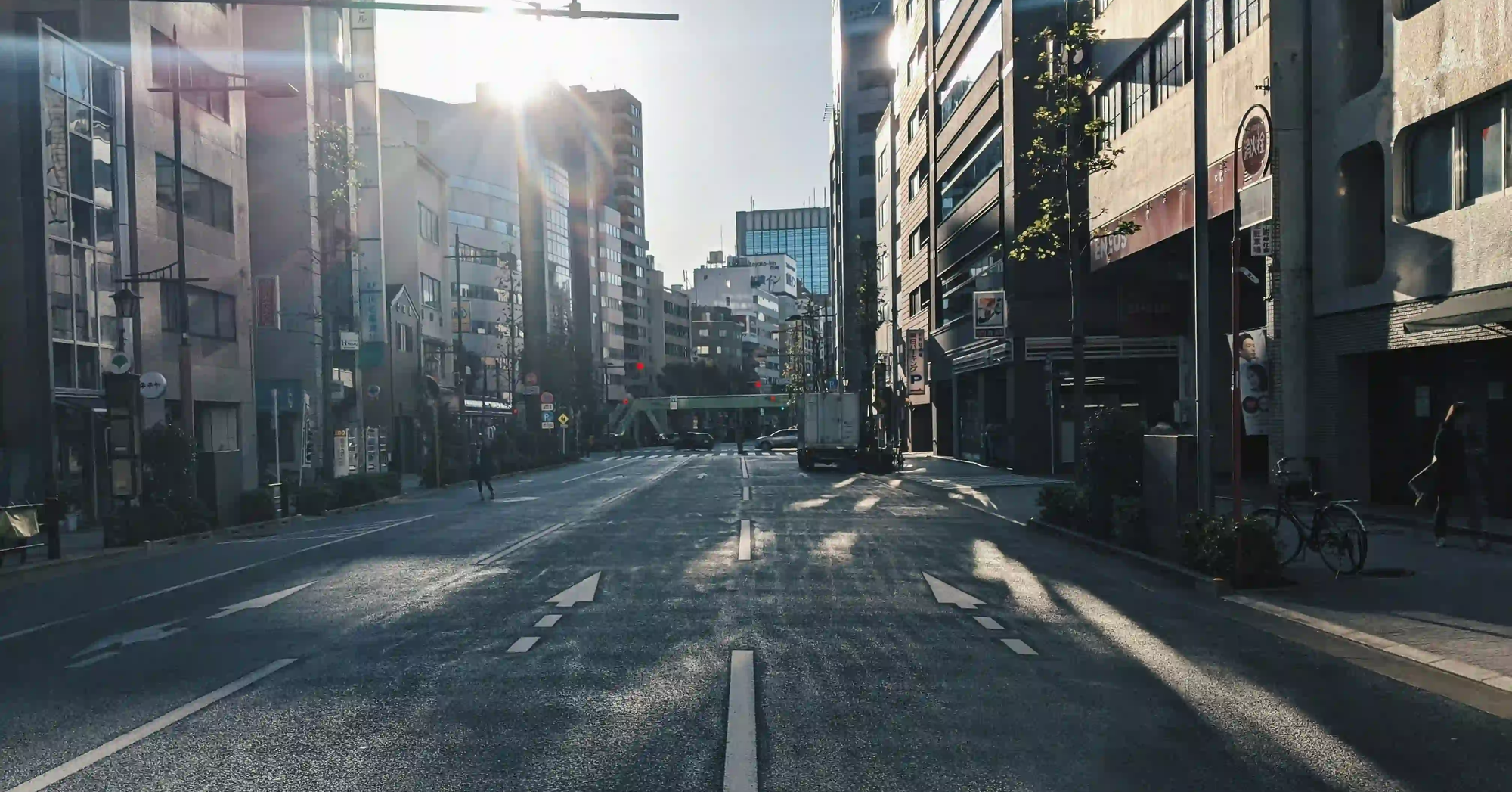 Bright sunlight flaring between tall buildings on a Japanese street, casting long, distinct shadows across the asphalt.