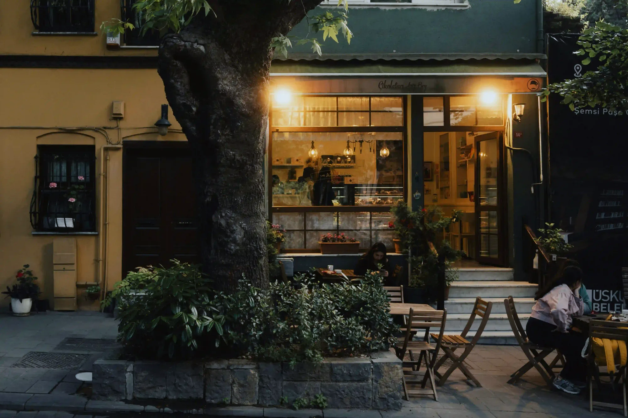 A warm, illuminated neighborhood cafe at dusk with a large tree in front and people sitting at wooden tables outside.