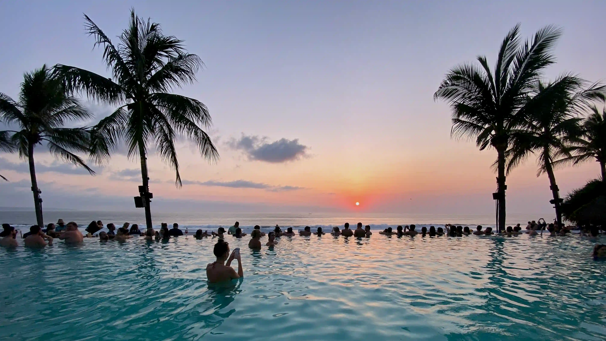 A crowded infinity pool in Bali at sunset, with numerous tourists holding smartphones, illustrating the mass tourism and content creation culture.