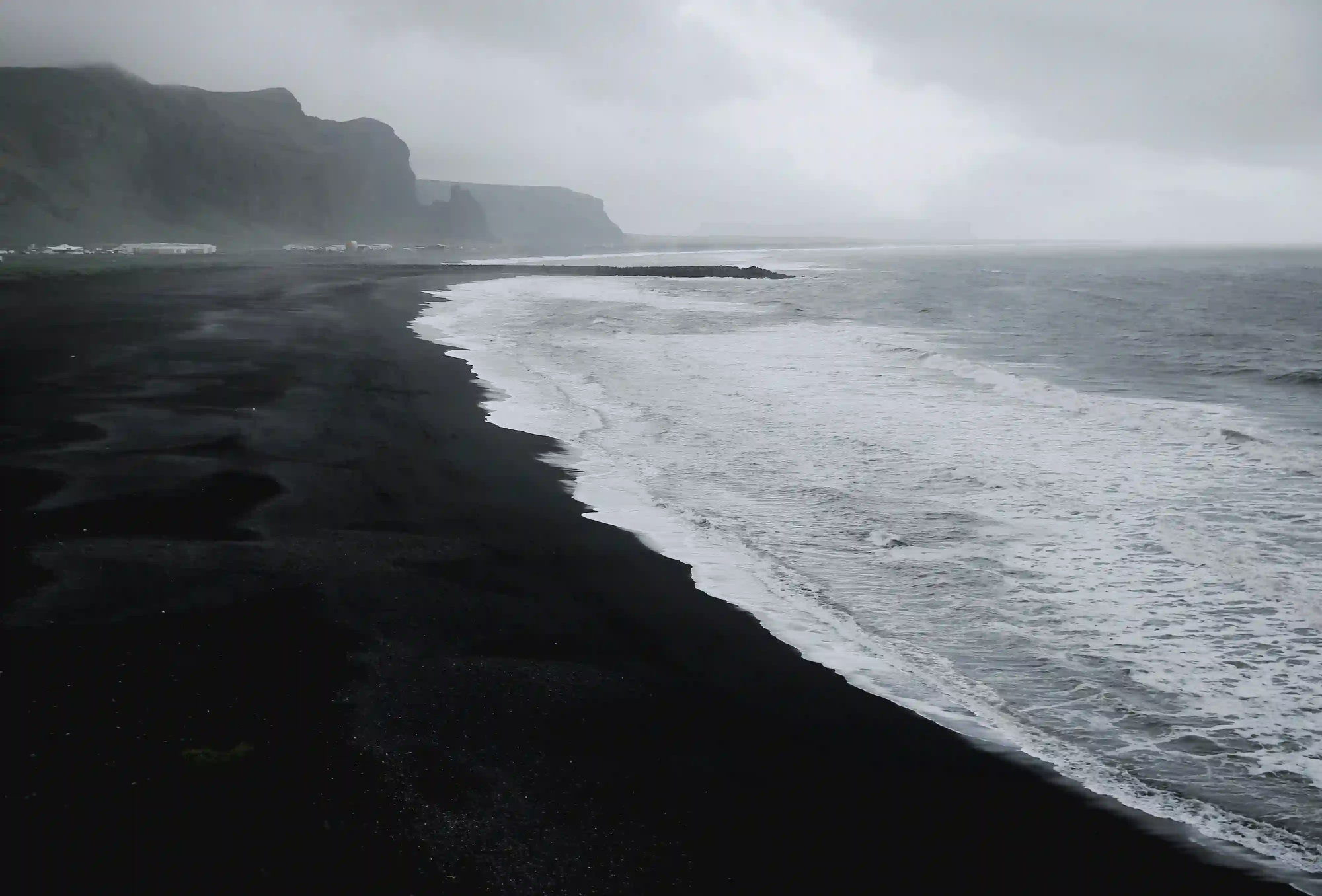 A dramatic and moody landscape of a black volcanic sand beach with crashing waves and dark cliffs under a foggy sky.