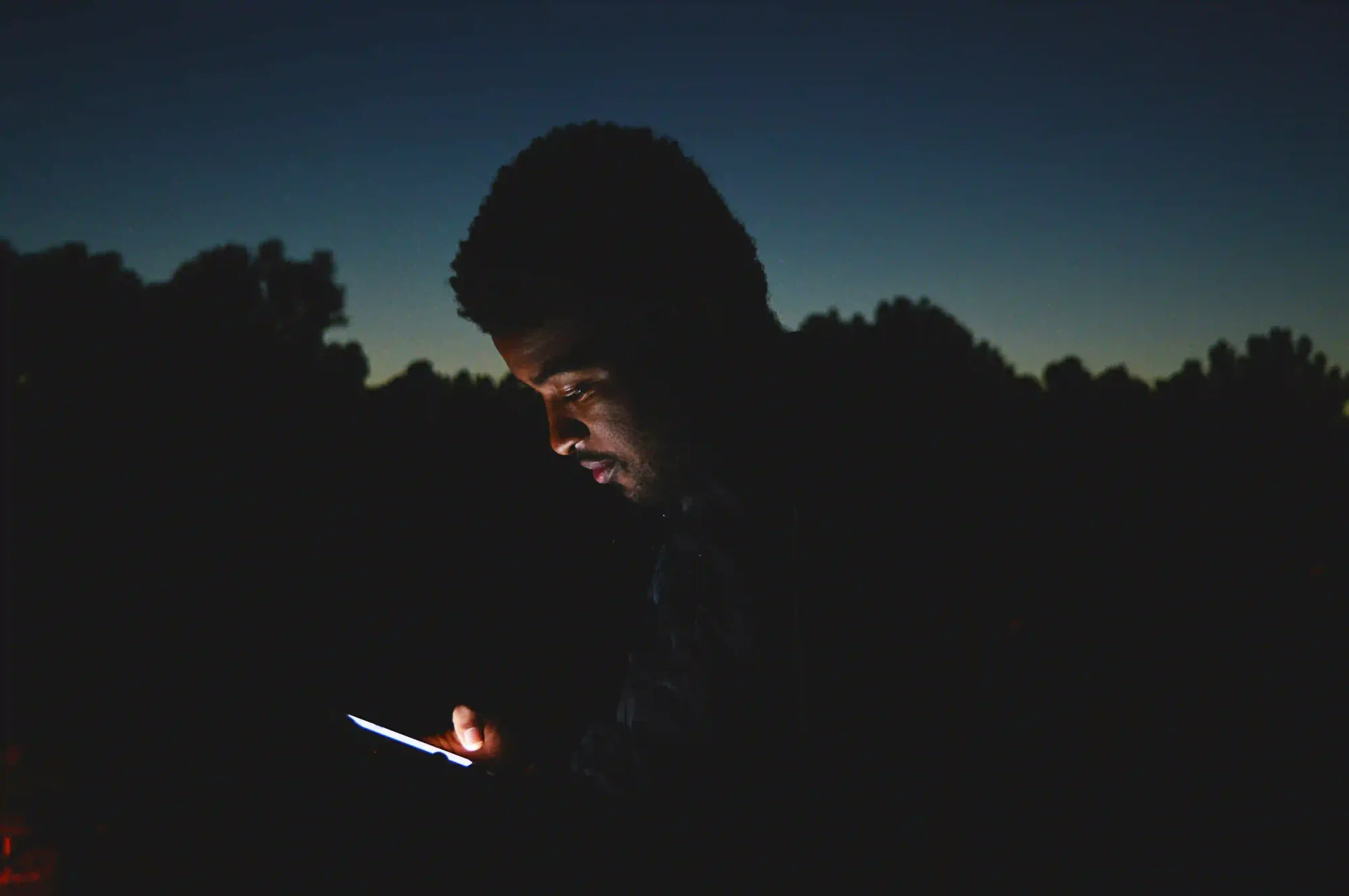 A person's face illuminated solely by a glowing smartphone screen in the dark, representing the artificial light that disrupts human dark adaptation and night vision.