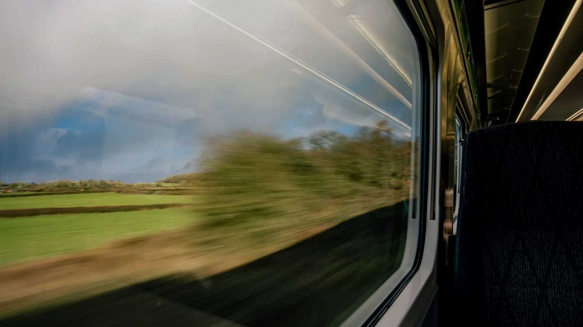A view from inside a moving train looking out the window at a blurred green landscape and cloudy sky, representing slow travel in Europe.