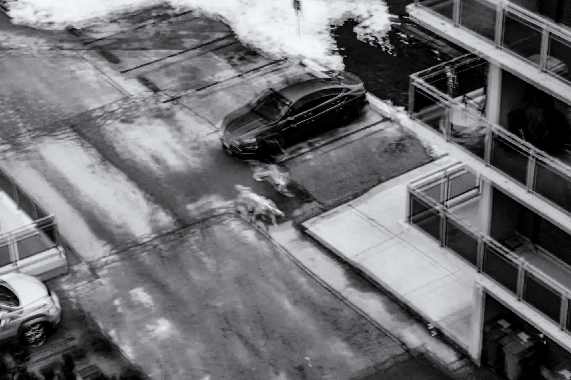 A black and white, slightly blurred aerial view of a paved street and modern apartment building, illustrating the cold and isolating nature of urban life.
