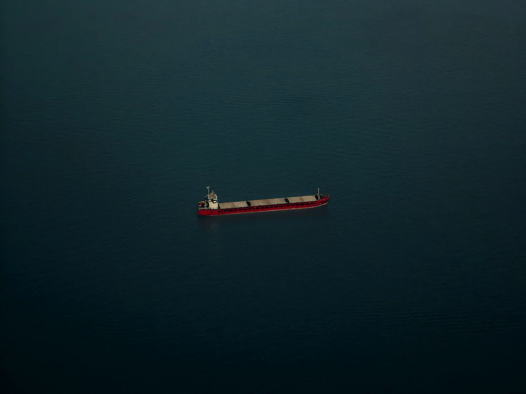 An aerial top-down view of a massive red cargo ship navigating through a dark, endless ocean, symbolizing global maritime trade.