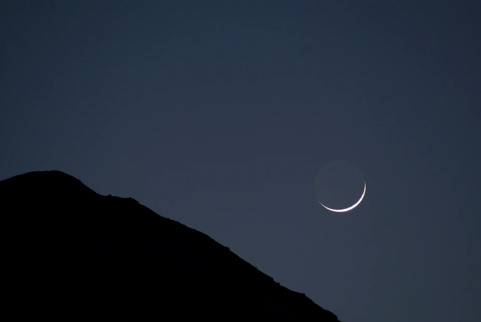 A minimalist dark mountain silhouette under a twilight sky with a thin crescent moon, representing the June 2026 planetary alignment.
