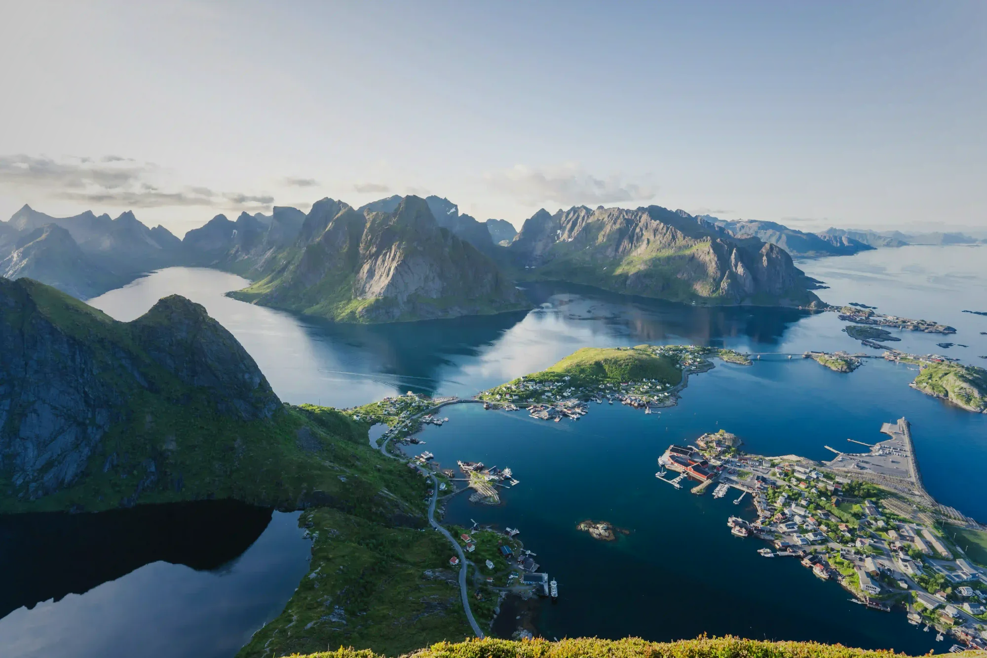 A breathtaking aerial view of the Lofoten Islands, Norway, showcasing dramatic green mountains plunging into deep blue fjords and a quiet coastal village.