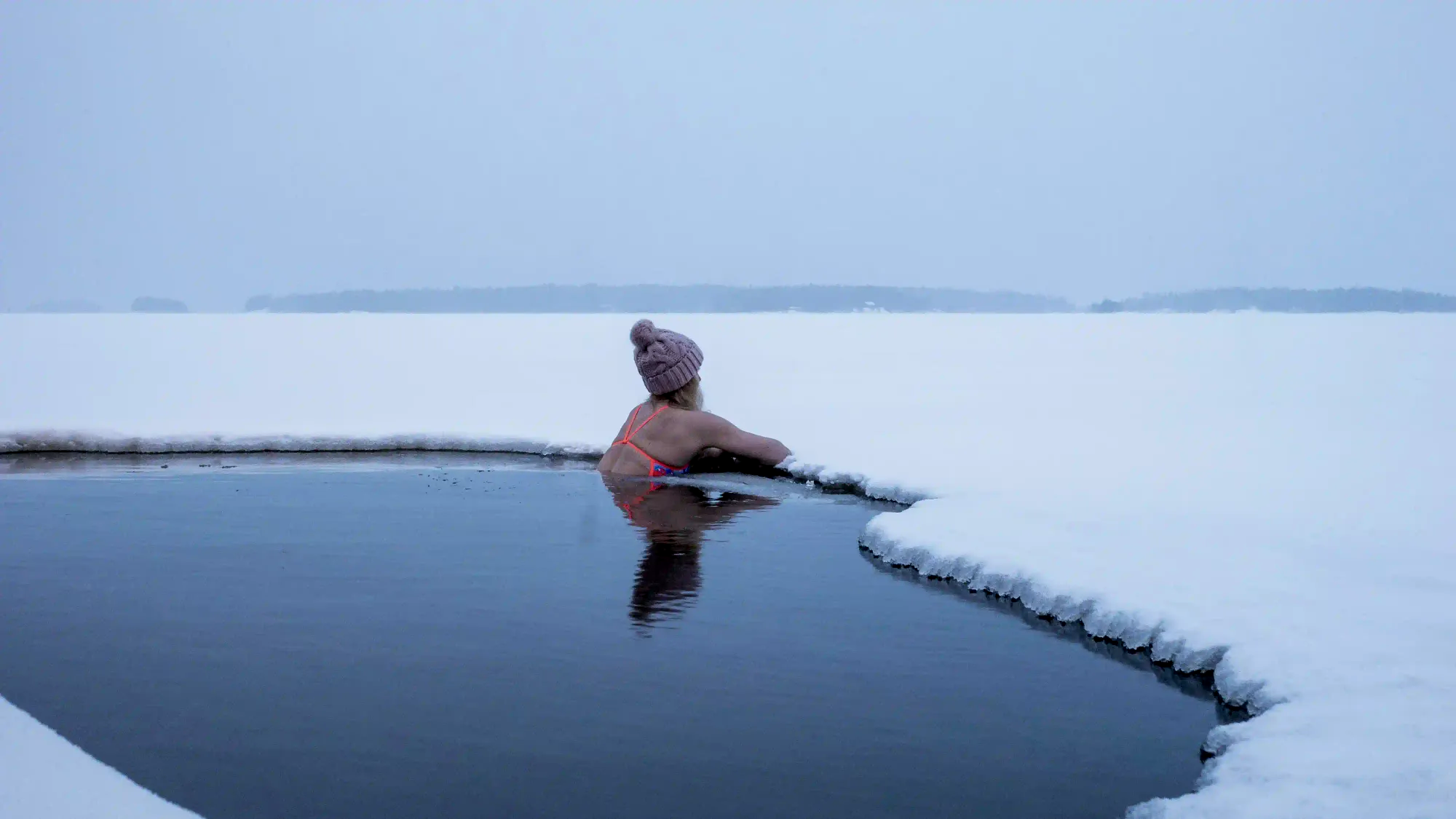 A person immersed in a dark avanto ice hole cut into a vast frozen lake, surrounded by a minimalist snow-covered landscape.
