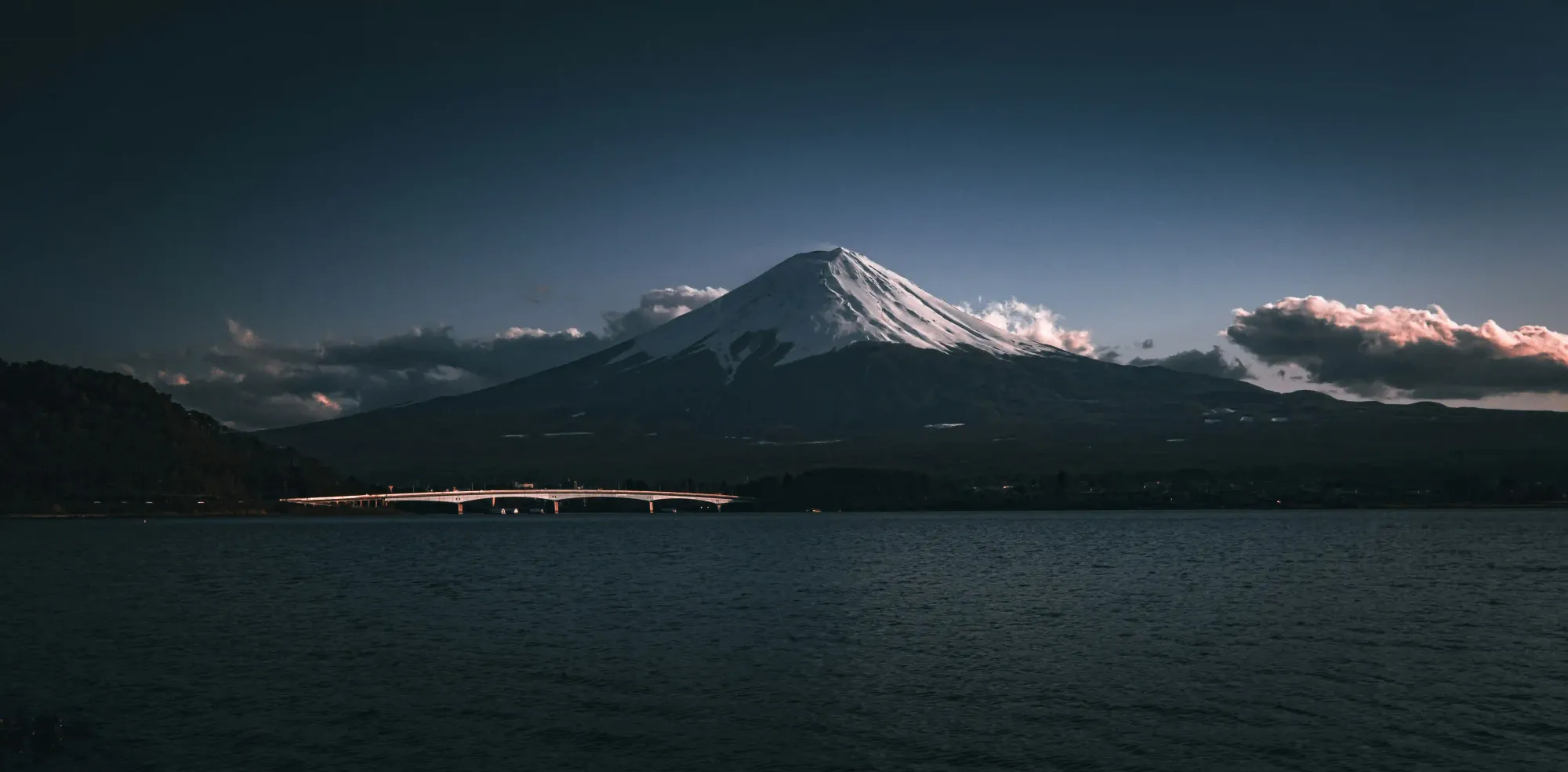 A moody, wide-angle landscape of Mount Fuji towering over a calm dark lake and a white bridge under a dramatic sky.