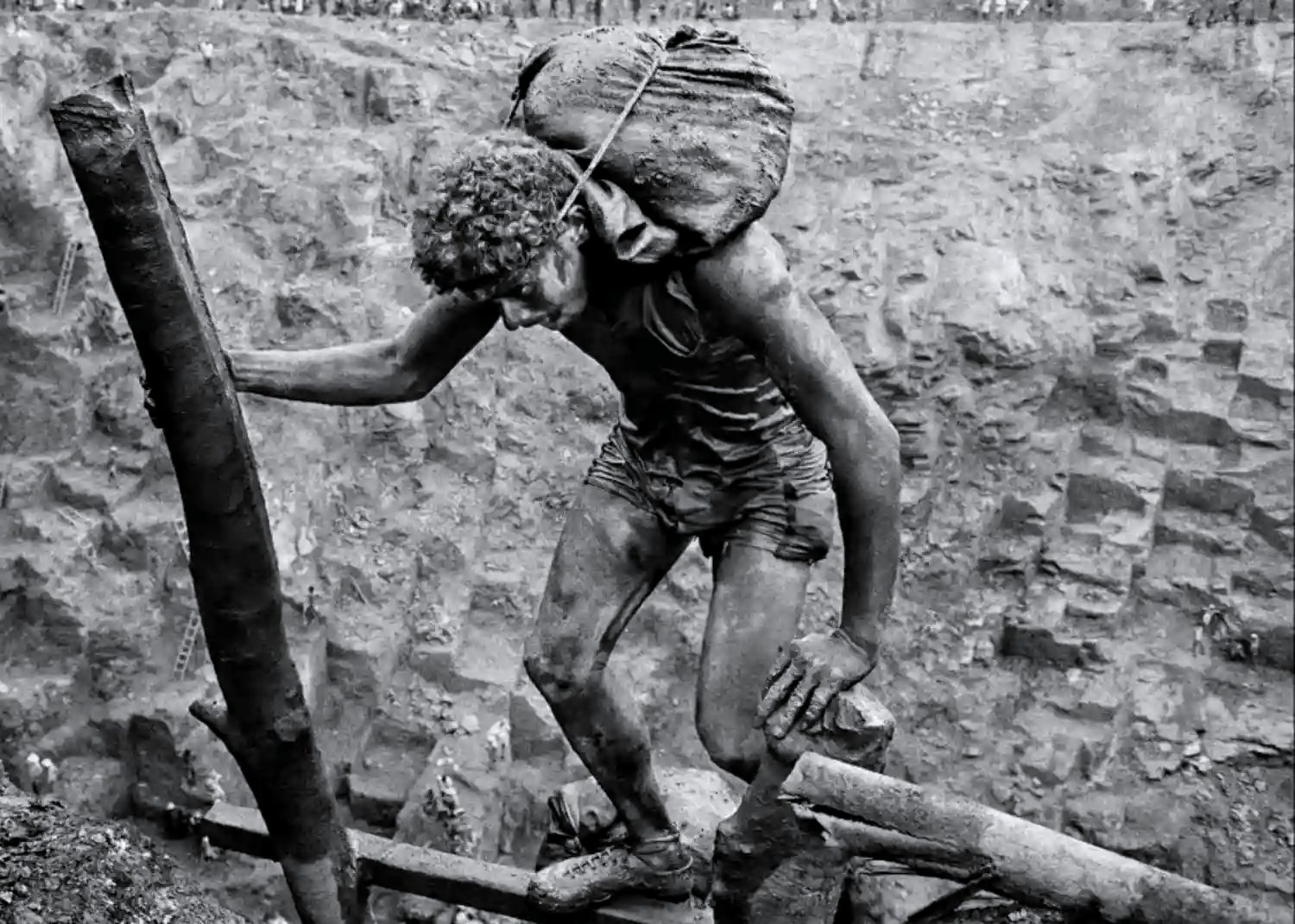 A powerful black and white photograph by Sebastião Salgado from the documentary The Salt of the Earth, depicting a mud-covered miner carrying a heavy sack on his back in a massive open-pit gold mine.