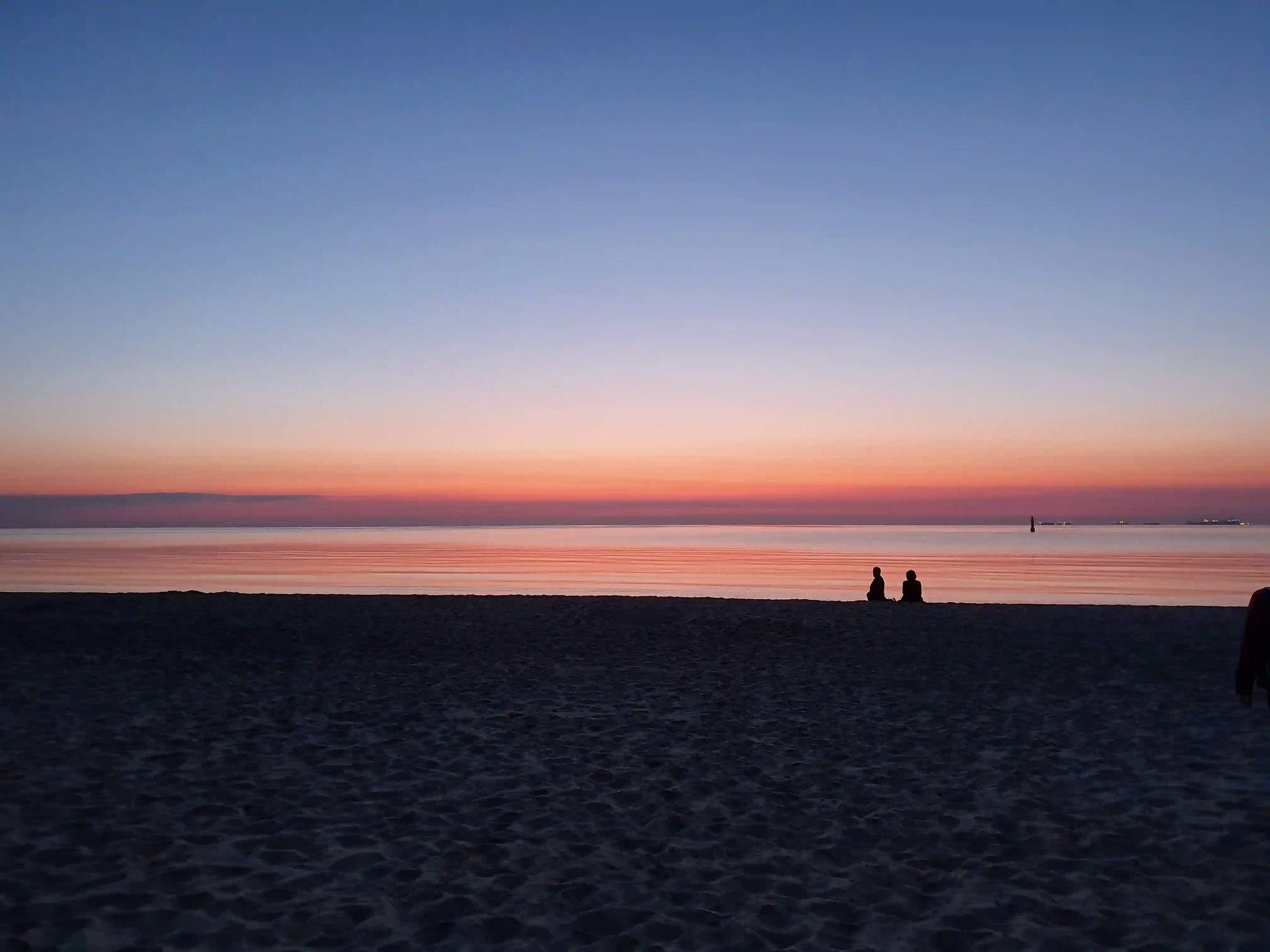 Two distant silhouettes sitting on a wide, tranquil beach at sunset in Sopot, Poland, gazing at the calm waters of the Baltic Sea under a pastel sky.