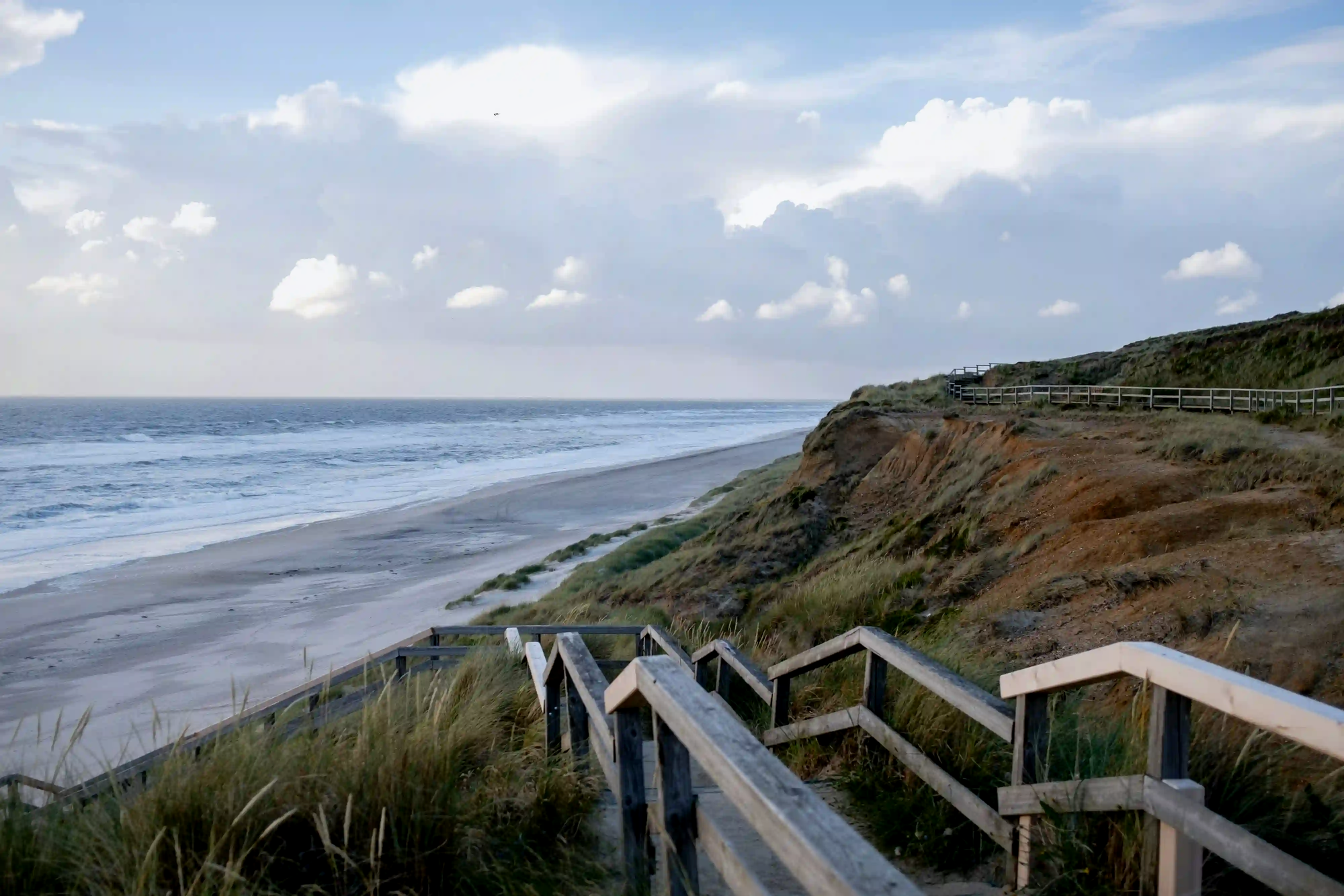 Wooden stairs leading down through the grassy dunes to the serene and empty beaches of Sylt Island, Germany, offering a cool North Sea breeze.