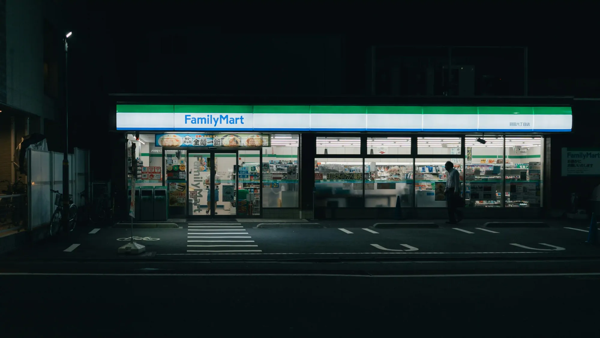 A brightly lit FamilyMart convenience store glowing on a dark, quiet street in Japan, representing the romanticization of mundane urban spaces.