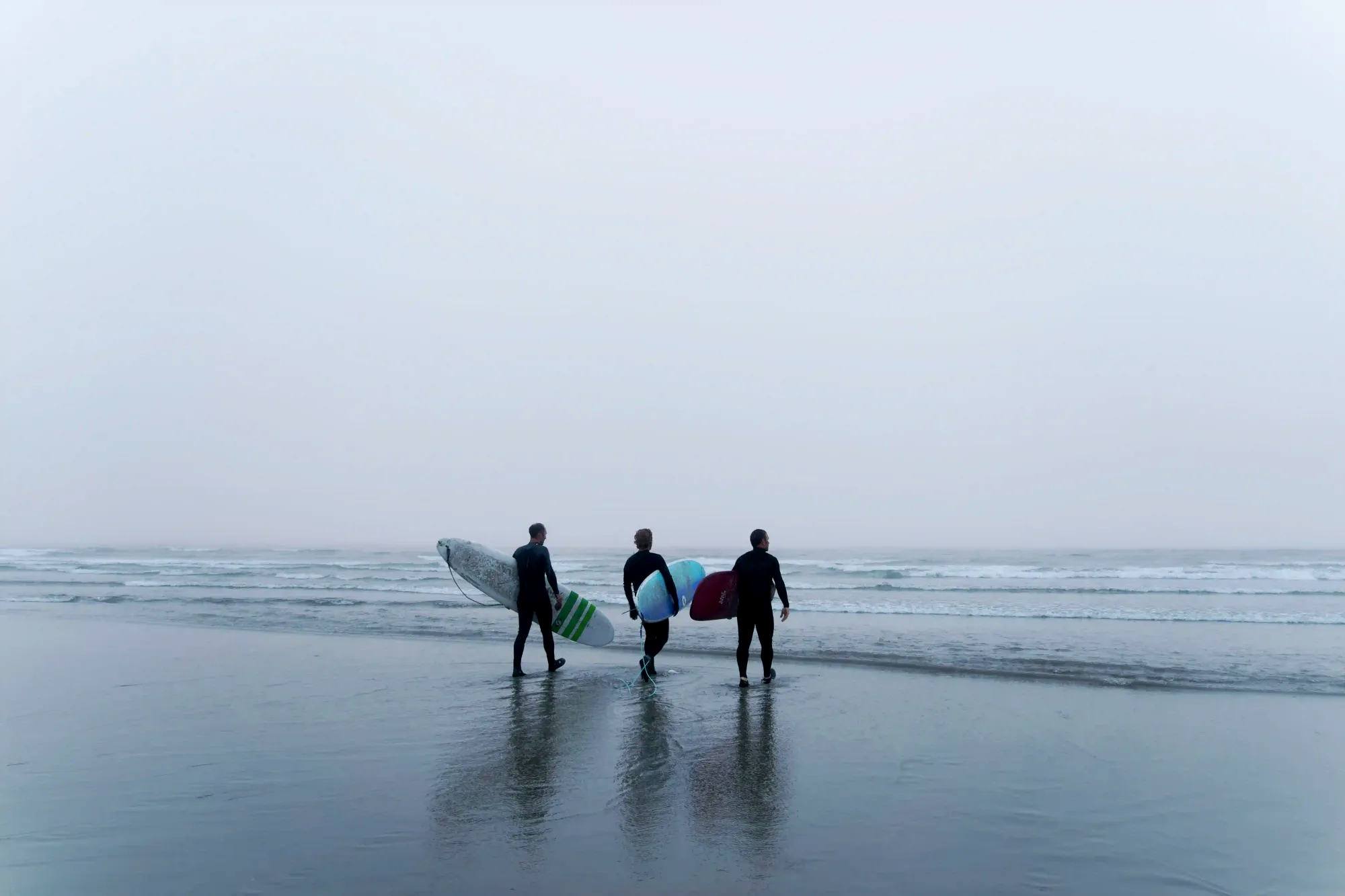 A cinematic view of three surfers in wetsuits walking towards the misty, cold waters of Tofino, Canada, highlighting the raw appeal of northern cold-water surfing.