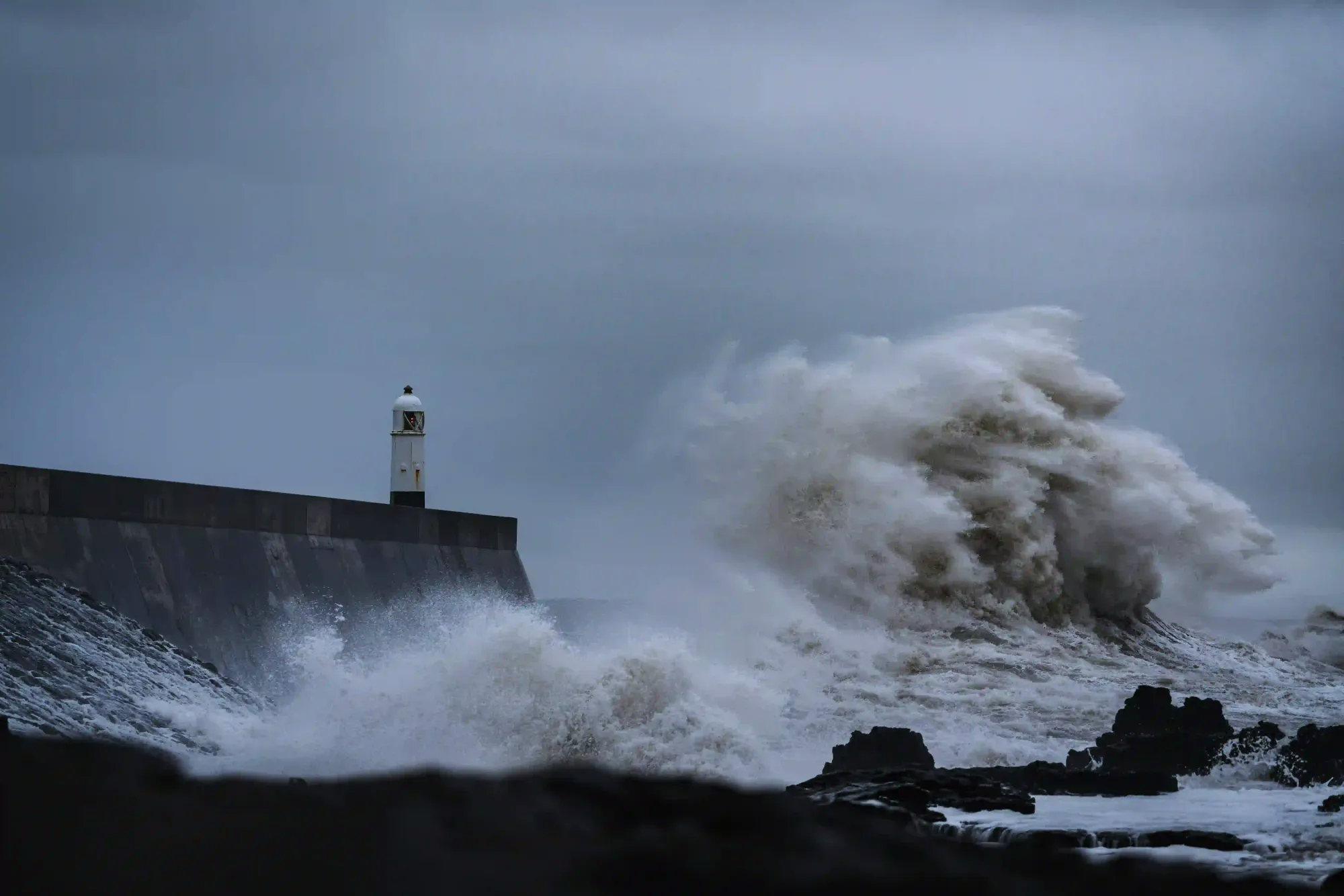 A massive ocean wave crashing violently against a dark concrete sea wall and a solitary lighthouse under a moody gray sky, representing harsh geographic isolation.
