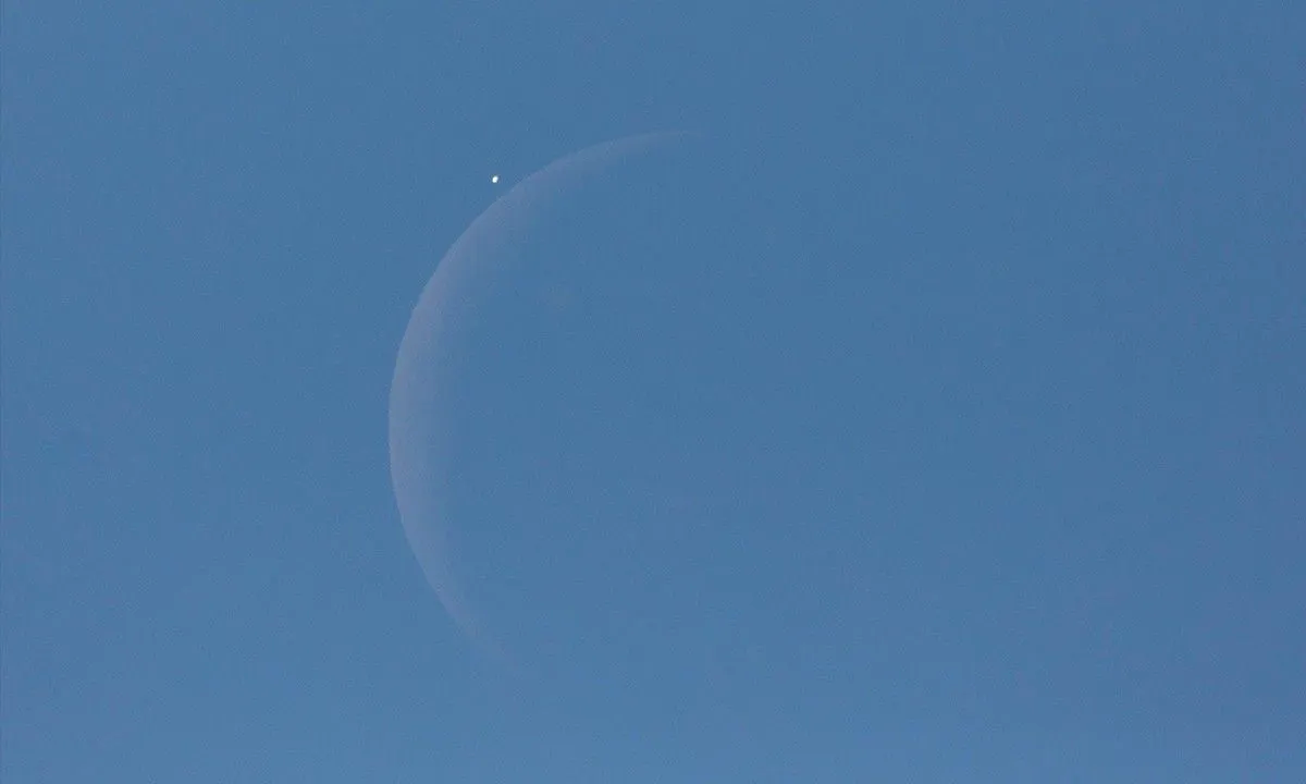 A raw telescopic view of a thin crescent moon about to eclipse the planet Venus against a blue daylight sky, illustrating a rare lunar occultation.