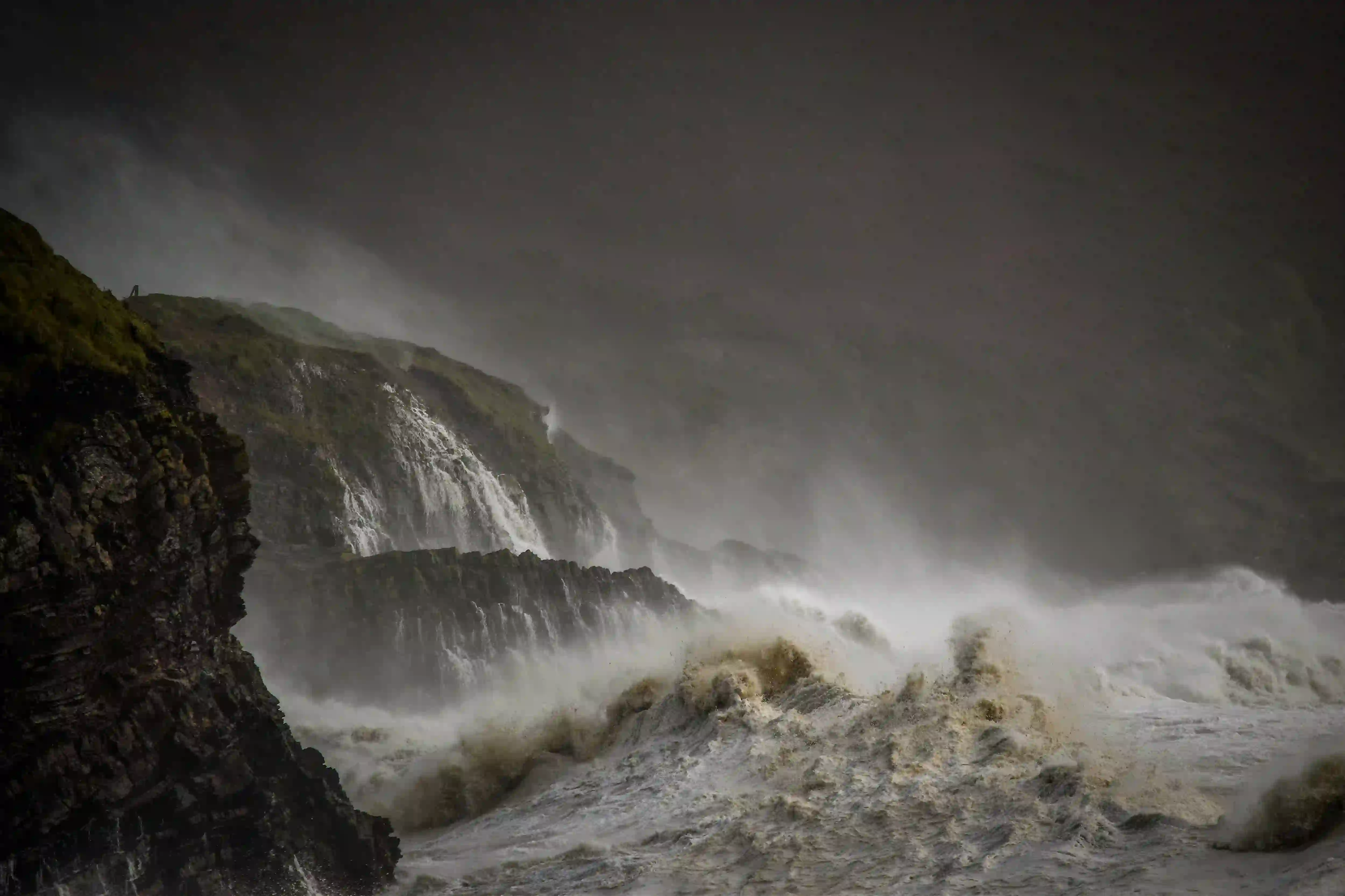 A moody, low-exposure photograph of violent, stormy ocean waves crashing heavily against dark, rugged coastal cliffs shrouded in mist.