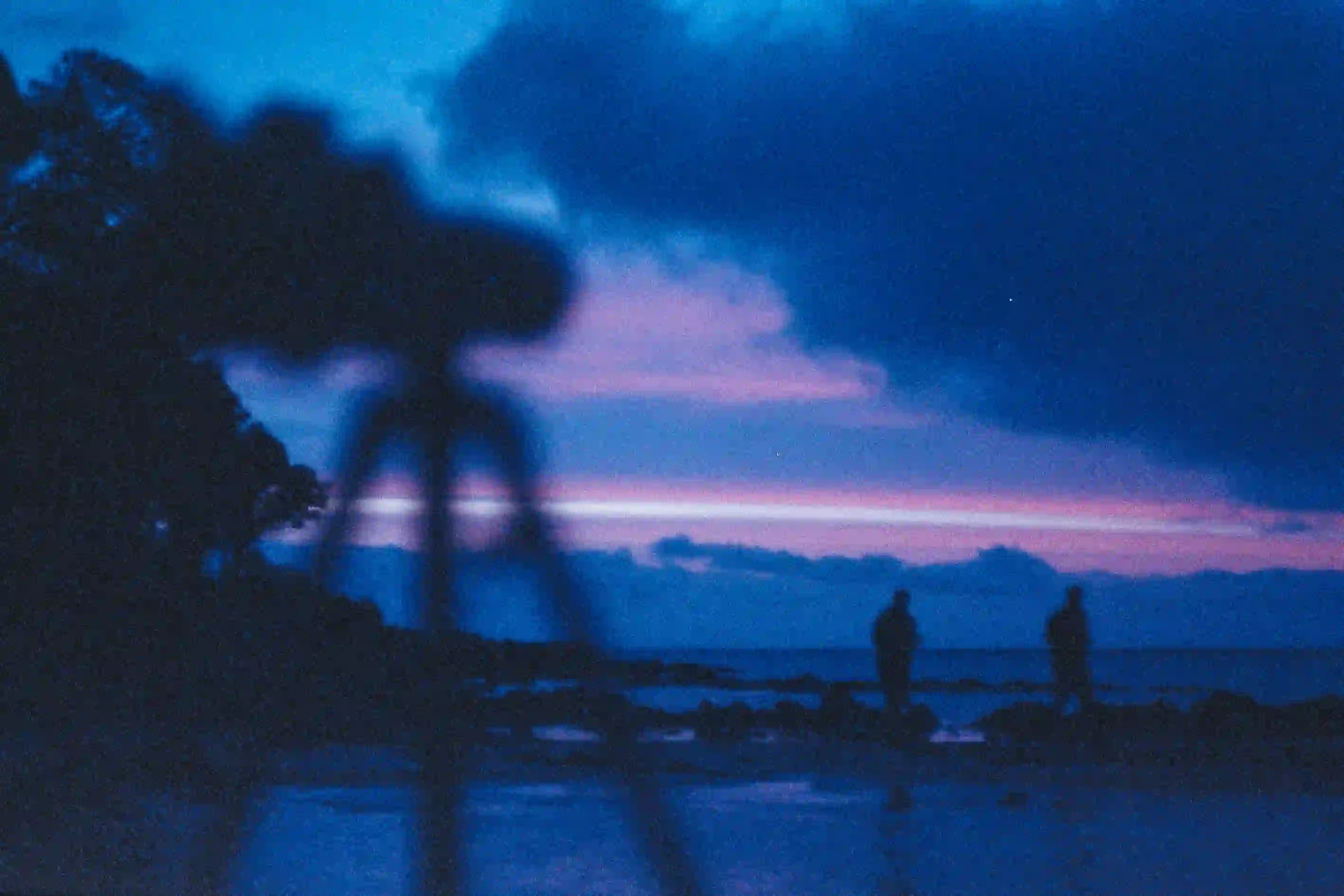 Blurred foreground of a camera tripod against a dark, moody blue-hour ocean sunset with two distant silhouetted figures.