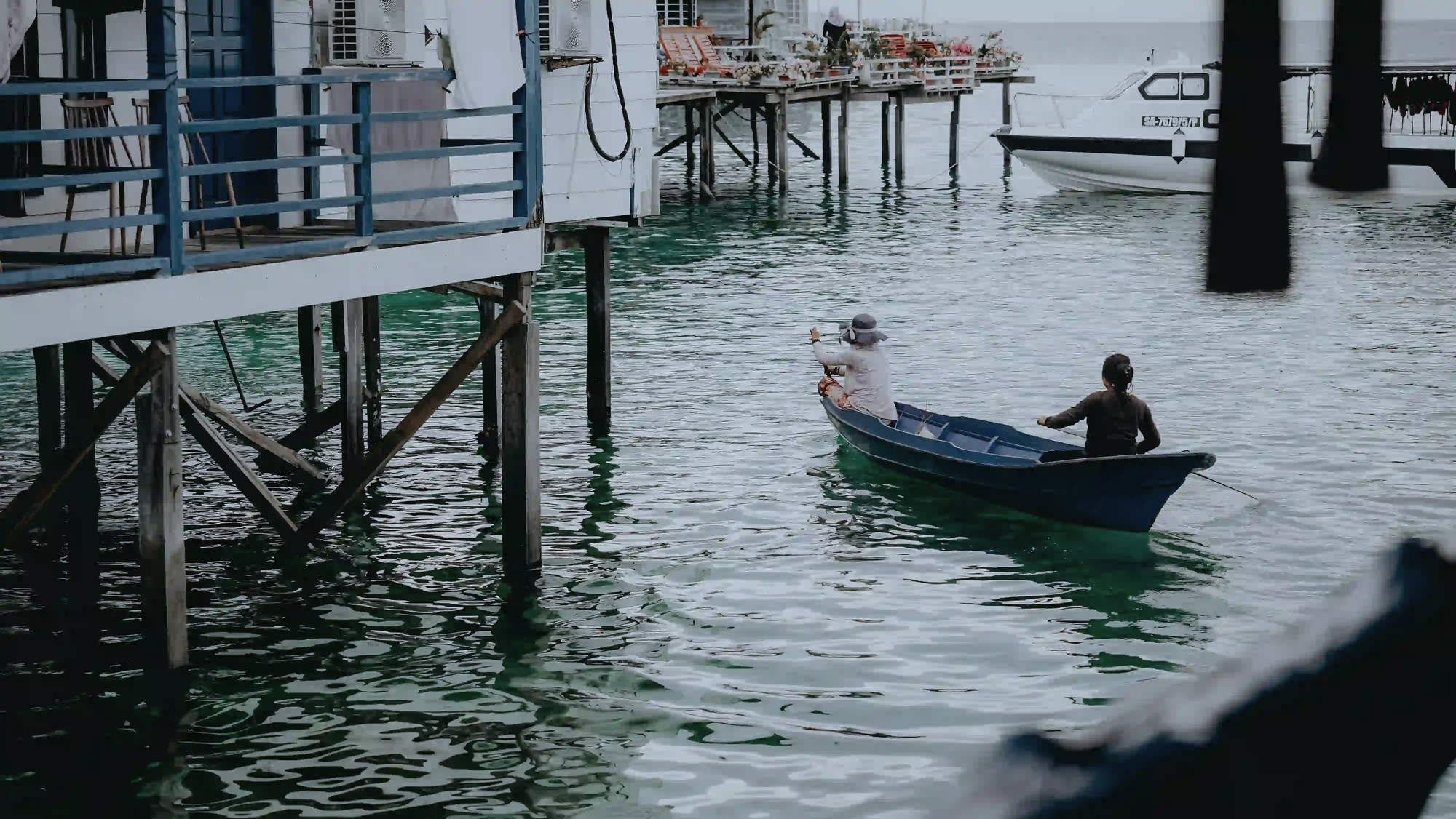 Two people rowing a small traditional wooden boat in clear waters, navigating past modern stilt houses equipped with air conditioning and a docked speedboat.