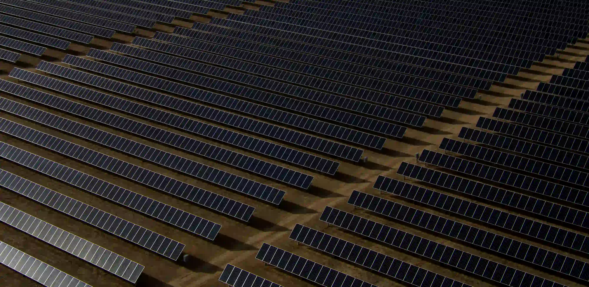 An aerial view of a massive solar panel farm, showing strict geometric lines and dark, low-exposure tones over bare earth.