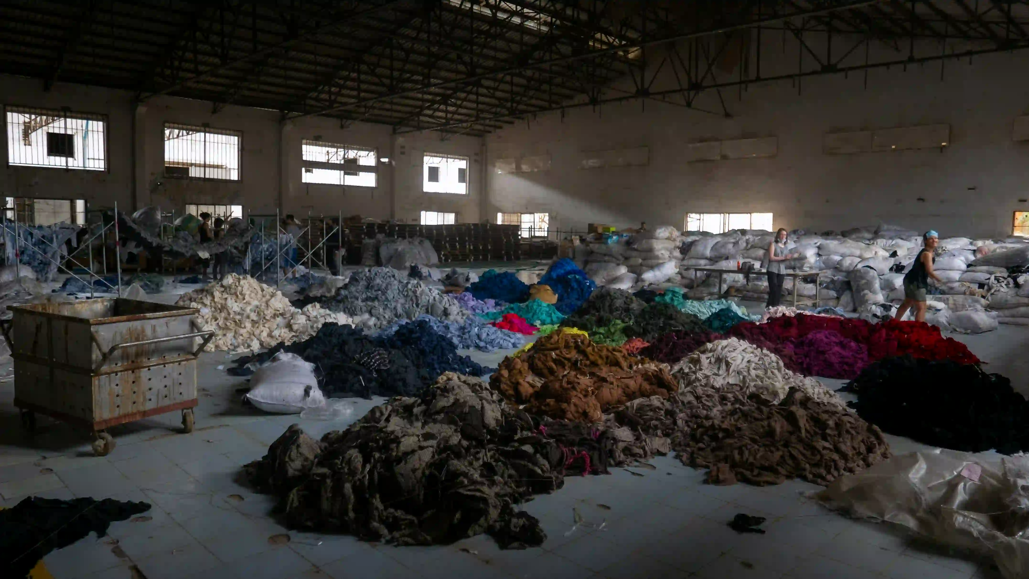 A dimly lit industrial warehouse floor covered in massive piles of sorted, discarded textile waste from the fast fashion industry.