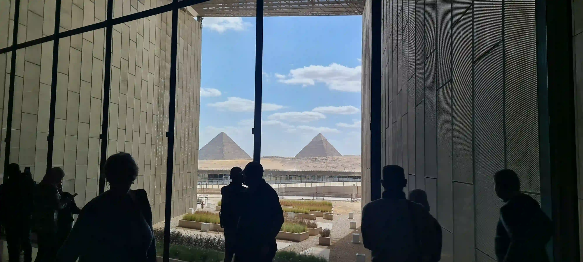 Silhouetted visitors inside the colossal, modern concrete structure of the Grand Egyptian Museum, looking out through massive glass windows at the ancient Pyramids of Giza on the horizon.