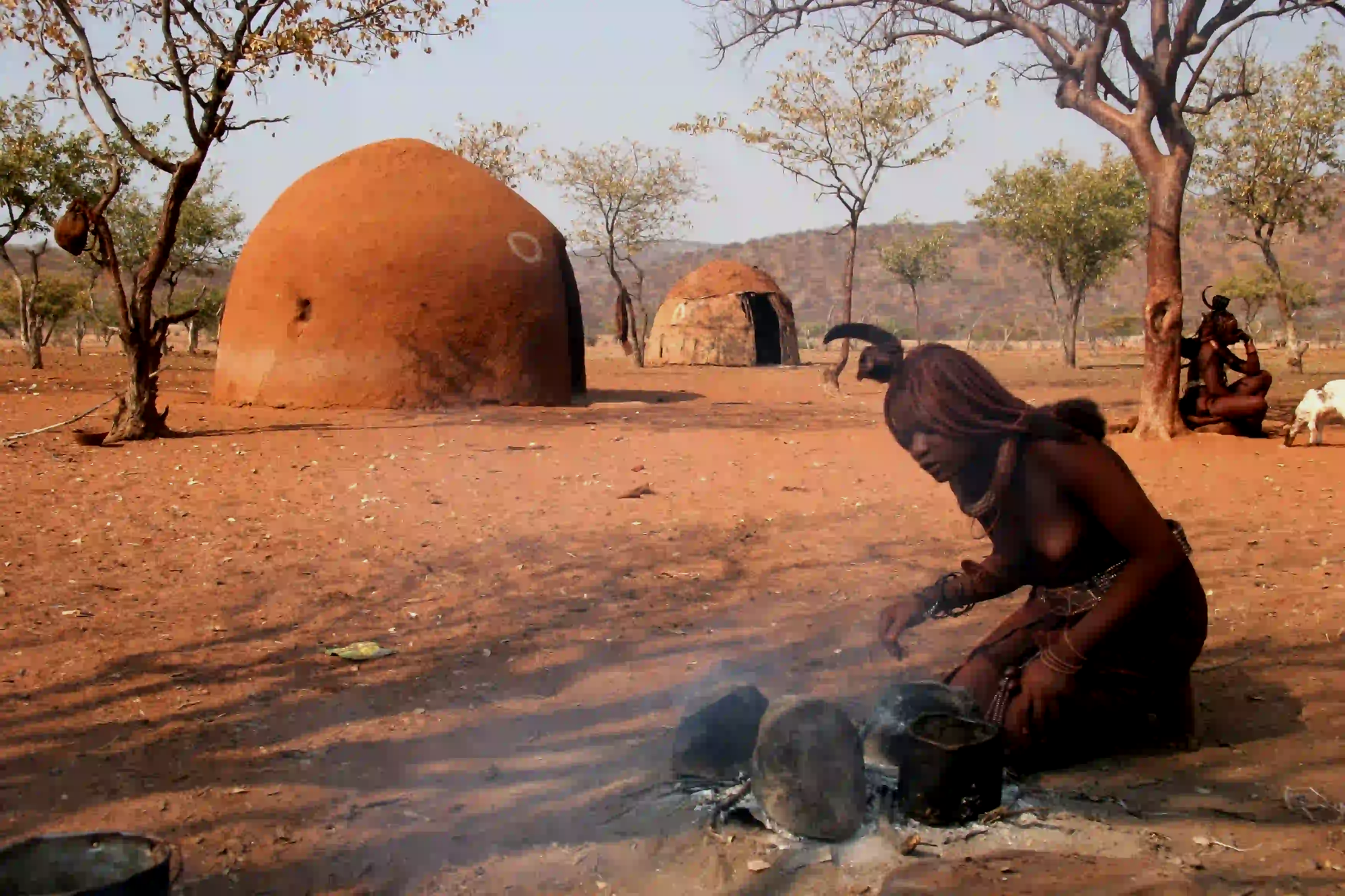 A Himba woman with traditional otjize-styled hair and skin tending a small fire in a dry Namibian landscape, with rounded earthen huts in the background.