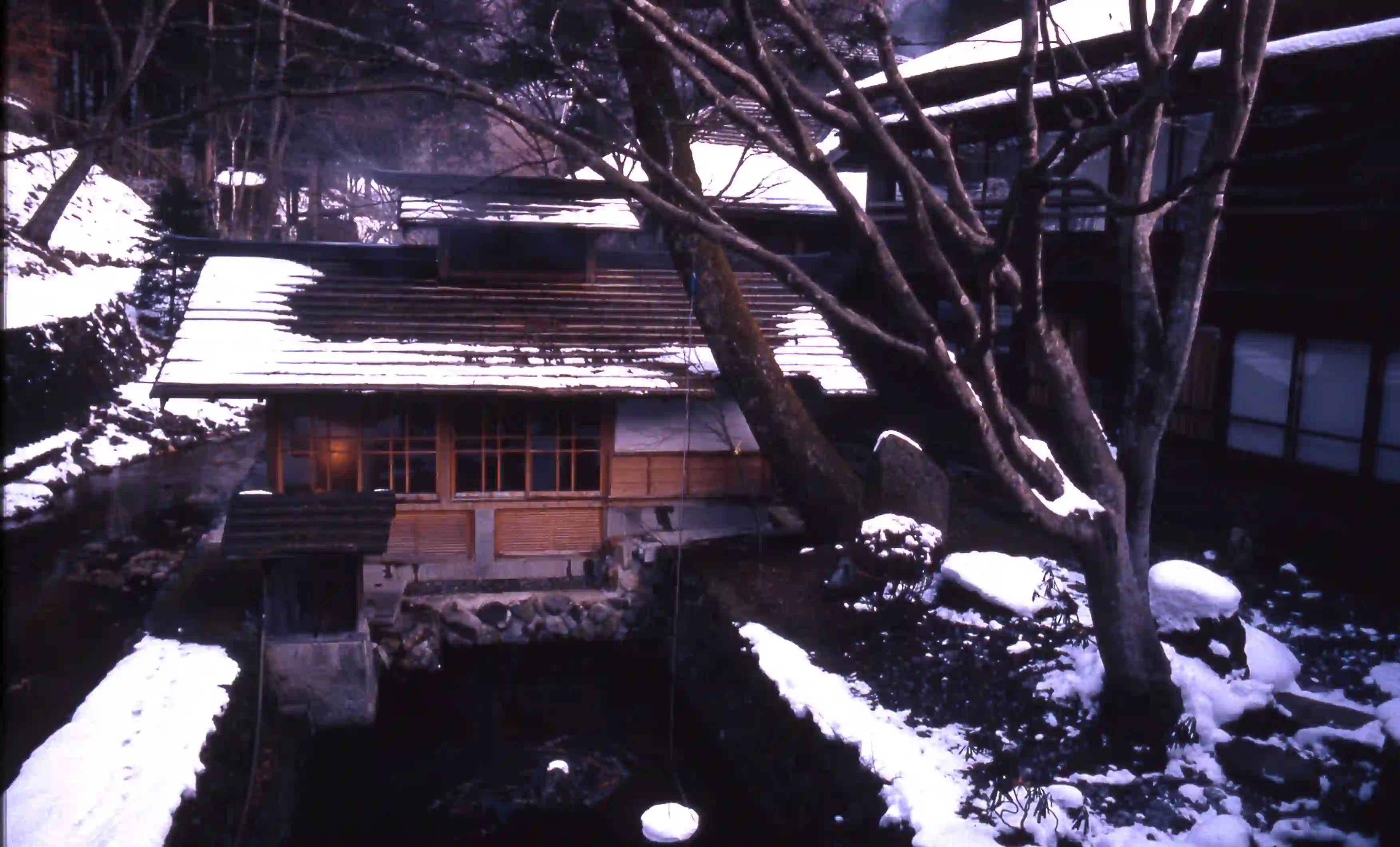 A traditional Japanese wooden bathhouse glowing with warm light in a dark, snow-covered winter landscape at Hōshi Ryokan.