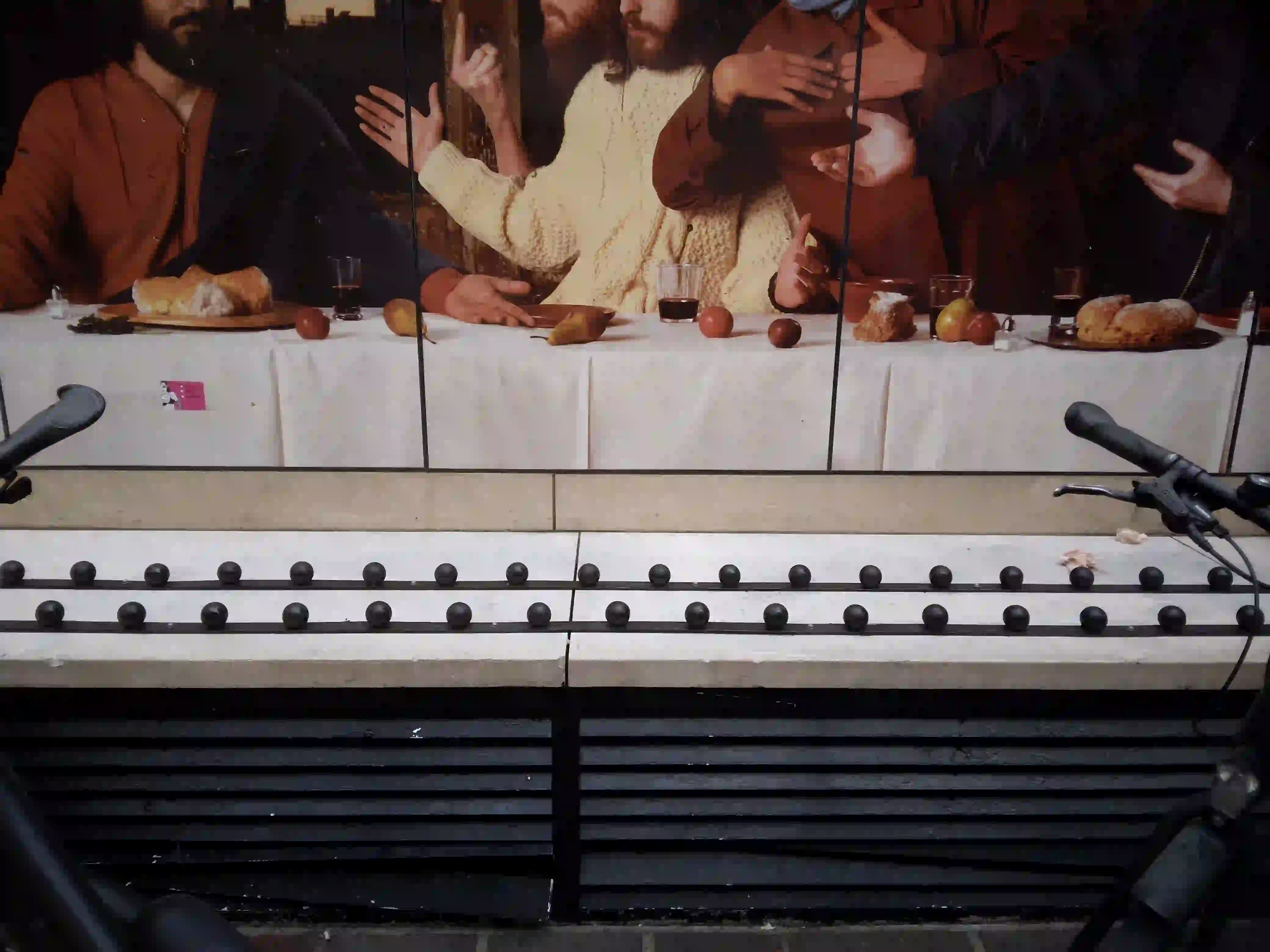 A stone ledge covered in anti-homeless metal studs, starkly juxtaposed against a background mural of The Last Supper in Dublin.