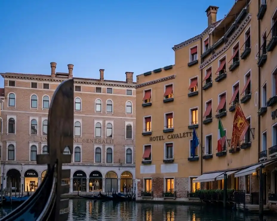 The historic yellow facade of Hotel Cavalletto e Doge Orseolo reflecting in a Venetian canal at dusk, viewed past the dark silhouette of a gondola prow.