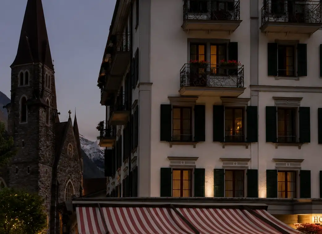 Warm light glows from the windows of a classic Swiss hotel at dusk, standing next to a historic stone church with snow-capped mountains in the background.