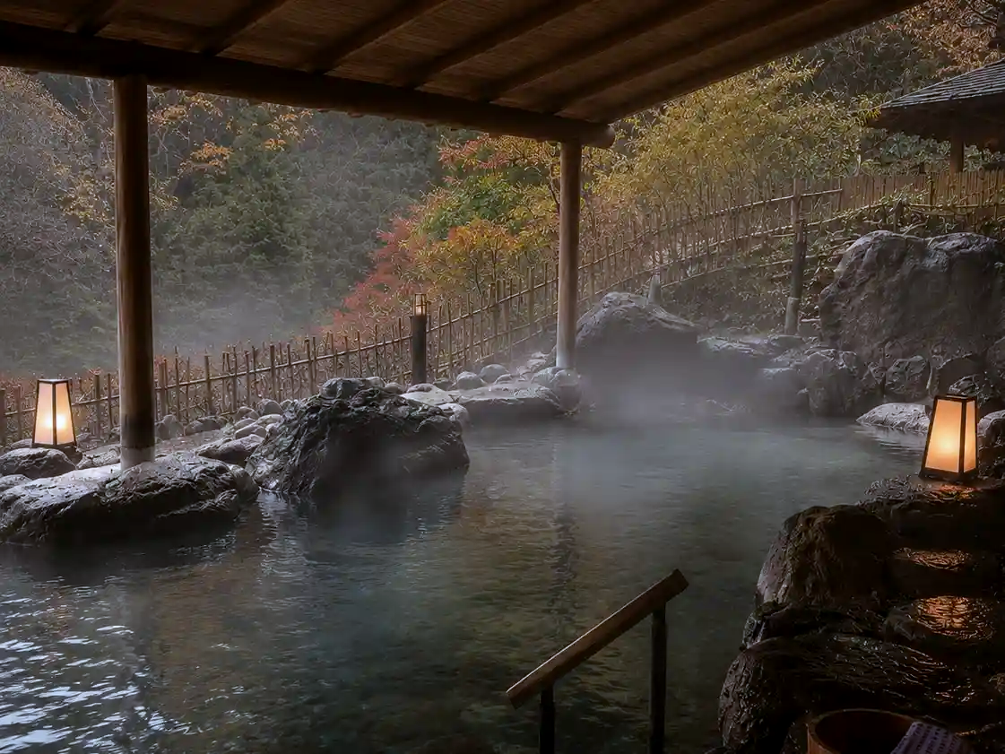 A detailed close-up of a steaming, open-air Japanese hot spring pool set among natural rocks. A wooden pavilion frames the scene, with two glowing paper lanterns. The background is a dense forest of evergreen and vibrant autumn-colored trees.