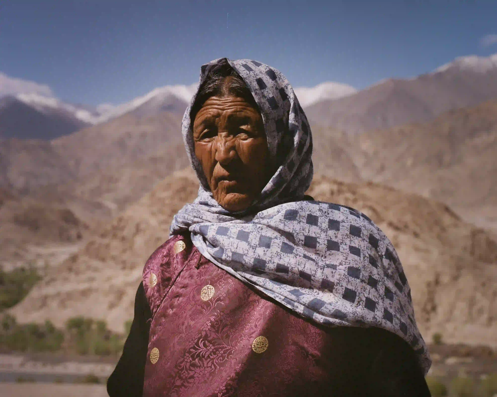 A portrait of an elderly Ladakhi woman with deeply weathered skin and traditional clothing, standing before the vast, barren mountains of the Himalayas.