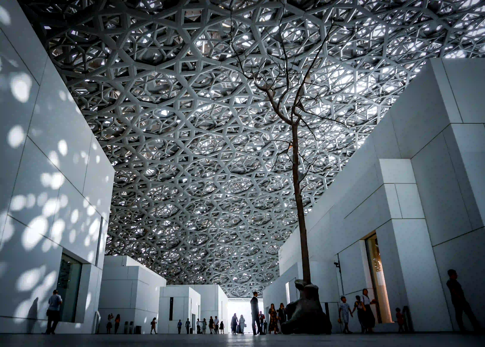 The massive, intricate steel dome of the Louvre Abu Dhabi filtering a mesmerizing rain of light onto white architectural walls, with silhouetted visitors walking below.