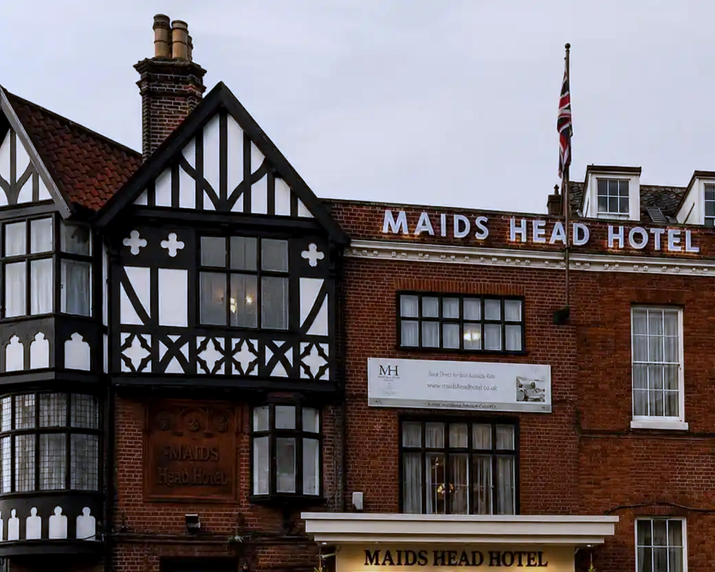 The exterior of the historic Maids Head Hotel, featuring a blend of traditional black-and-white half-timbered architecture and red brick, topped with an illuminated sign and a British flag.