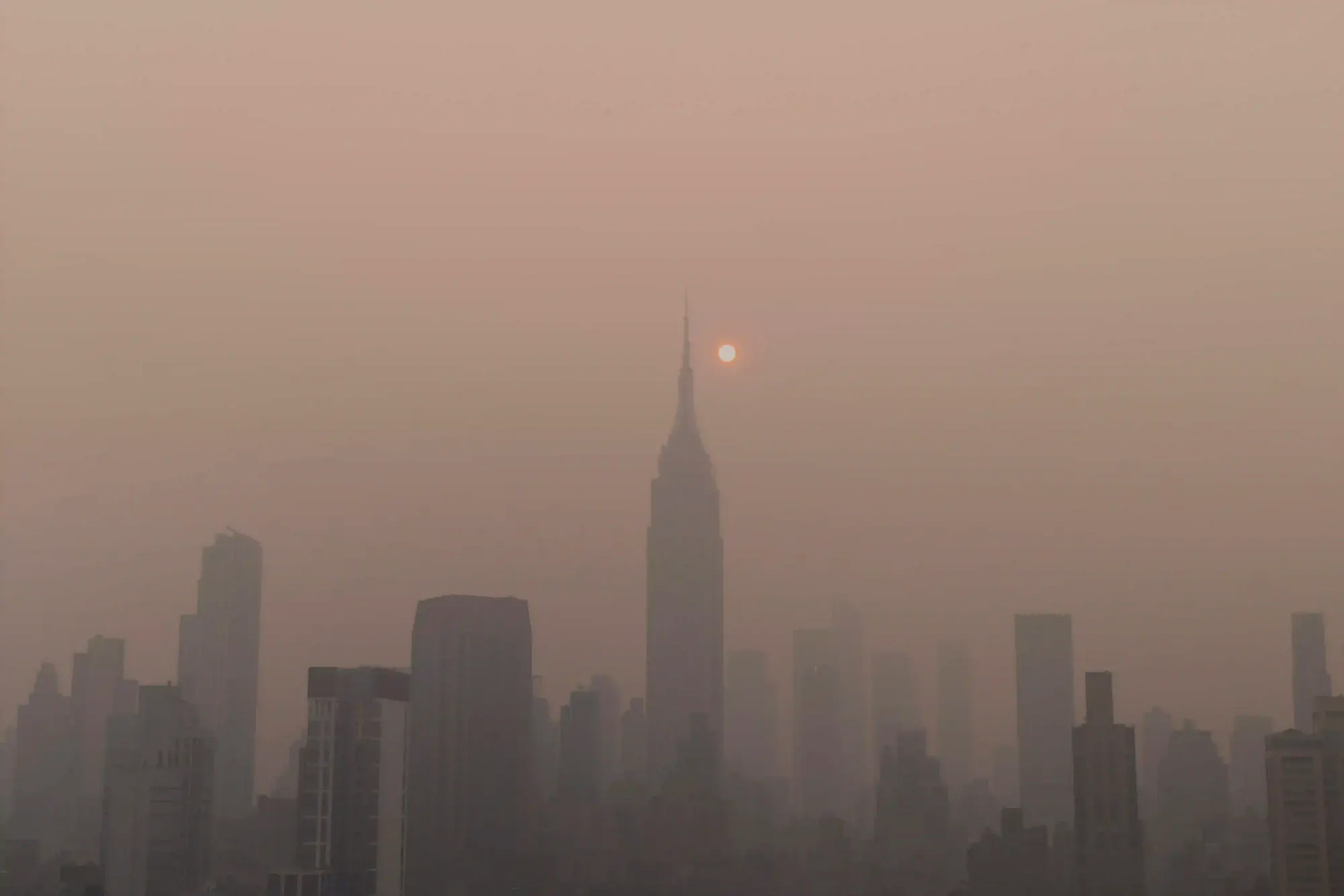 The New York City skyline and Empire State Building completely engulfed in a thick, apocalyptic orange haze from distant wildfire smoke.