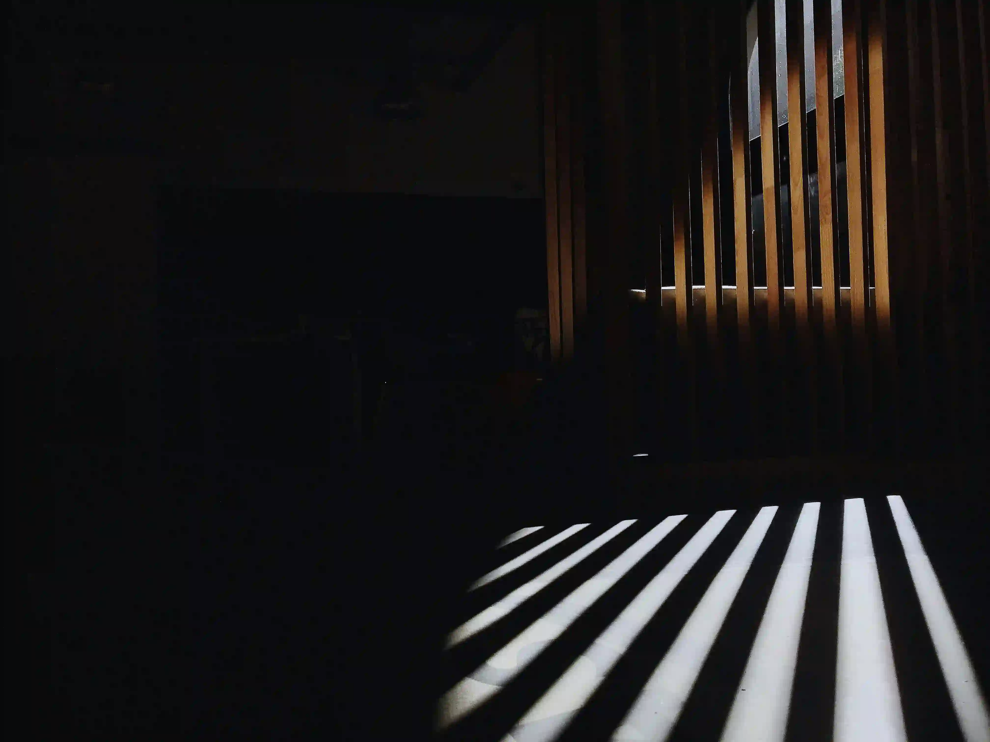 Abstract play of light and shadow through wooden slats, evoking a sense of solitude and quiet interiors.