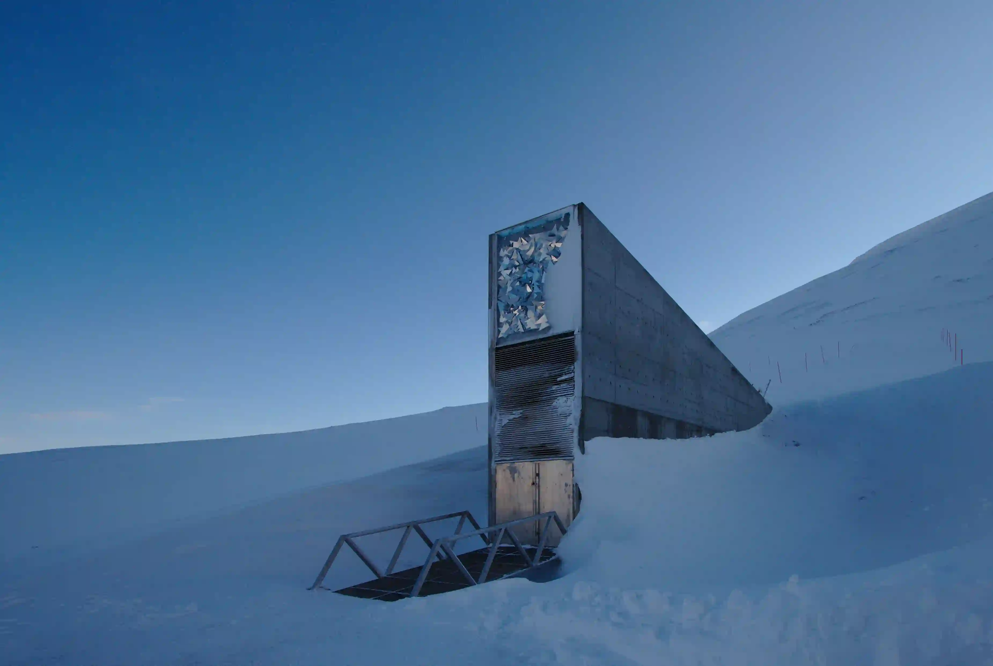 The sharp concrete entrance of the Svalbard Global Seed Vault jutting out from a snow-covered mountain under a cold blue Arctic sky.