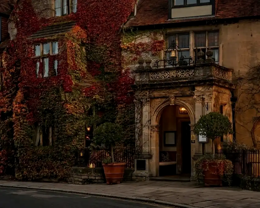 The historic stone facade of The Old Bell Hotel in England, heavily draped in autumn ivy with an ornate arched entrance and warm glowing lights.