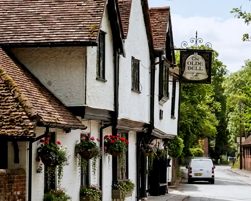 A daytime exterior view of The Olde Bell, featuring a historic white building with hanging flower baskets, gabled roofs, and a traditional hanging sign dating back to 1135.