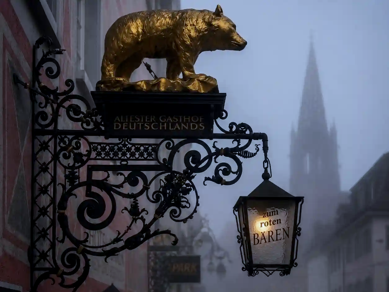An ornate wrought-iron sign featuring a golden bear and a glowing lantern reading "zum roten BÄREN", set against a foggy backdrop of a gothic cathedral spire.