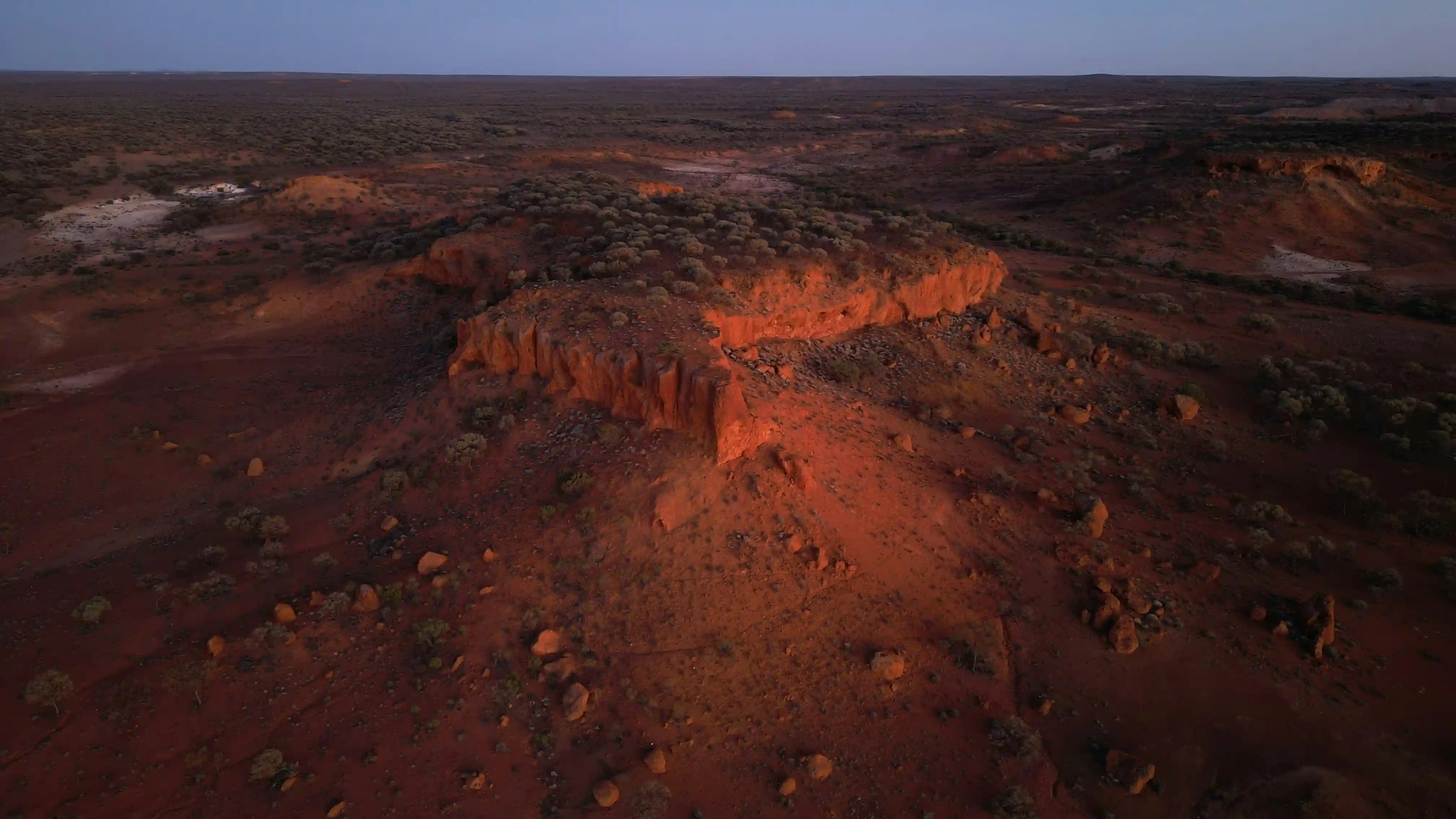 Aerial view of a vast, shadowed red dirt landscape in the Australian outback at dusk, representing the geographical terrain mapped by ancient Aboriginal Songlines.