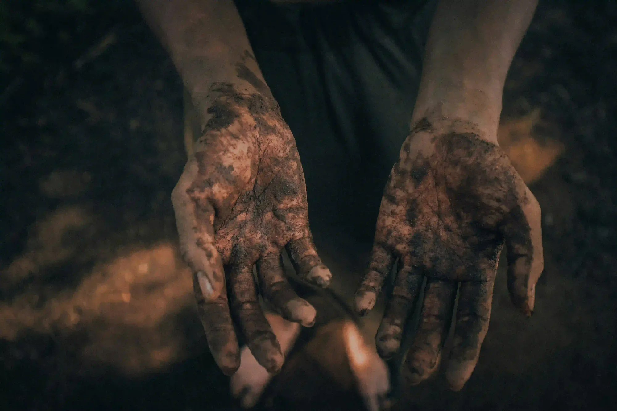 A moody, close-up shot of two hands deeply covered in dark, wet soil, representing the raw physical reality of working with the earth.