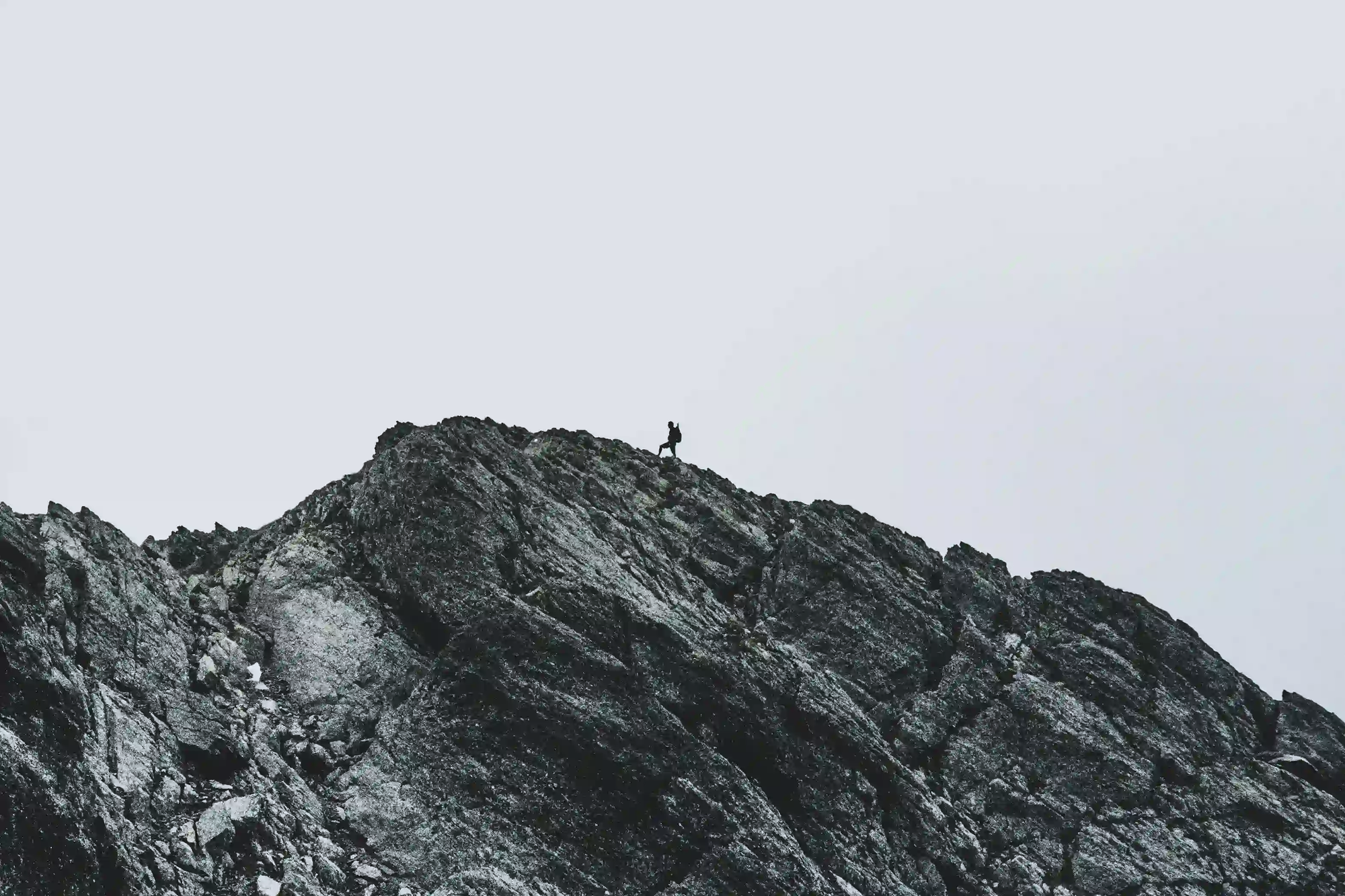 A tiny human silhouette standing on a massive, dark mountain ridge against a vast minimalist white sky, highlighting the overwhelming scale of nature.